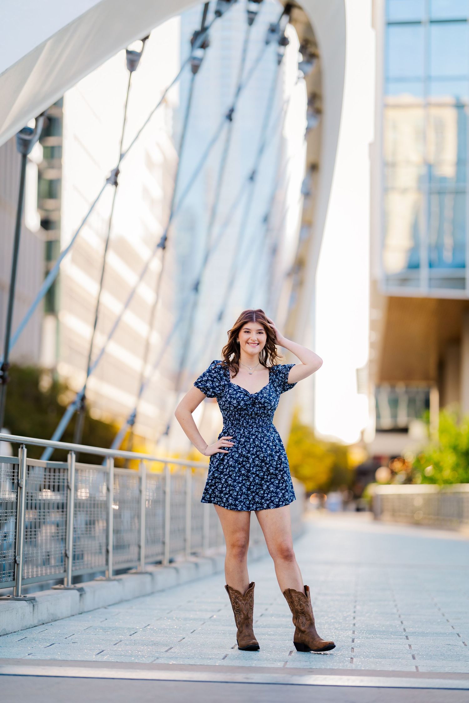 Young woman in blue floral dress and cowboy boots posing under modern bridge structure.