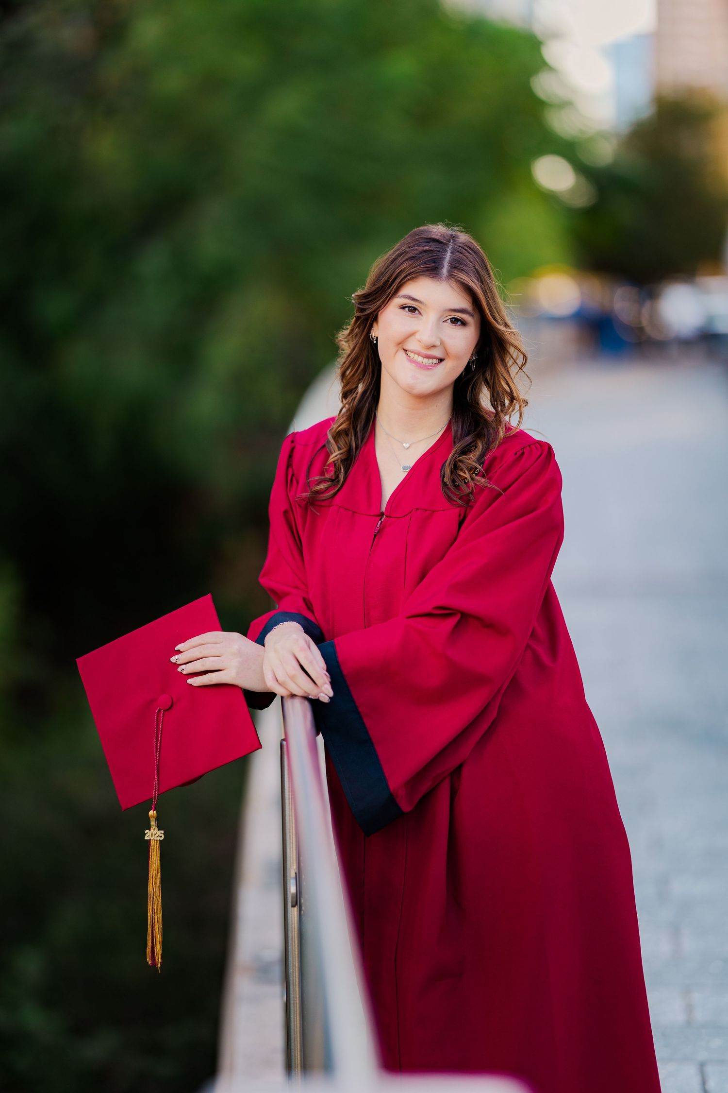Smiling female graduate in red cap and gown leaning on railing for outdoor graduation portrait.