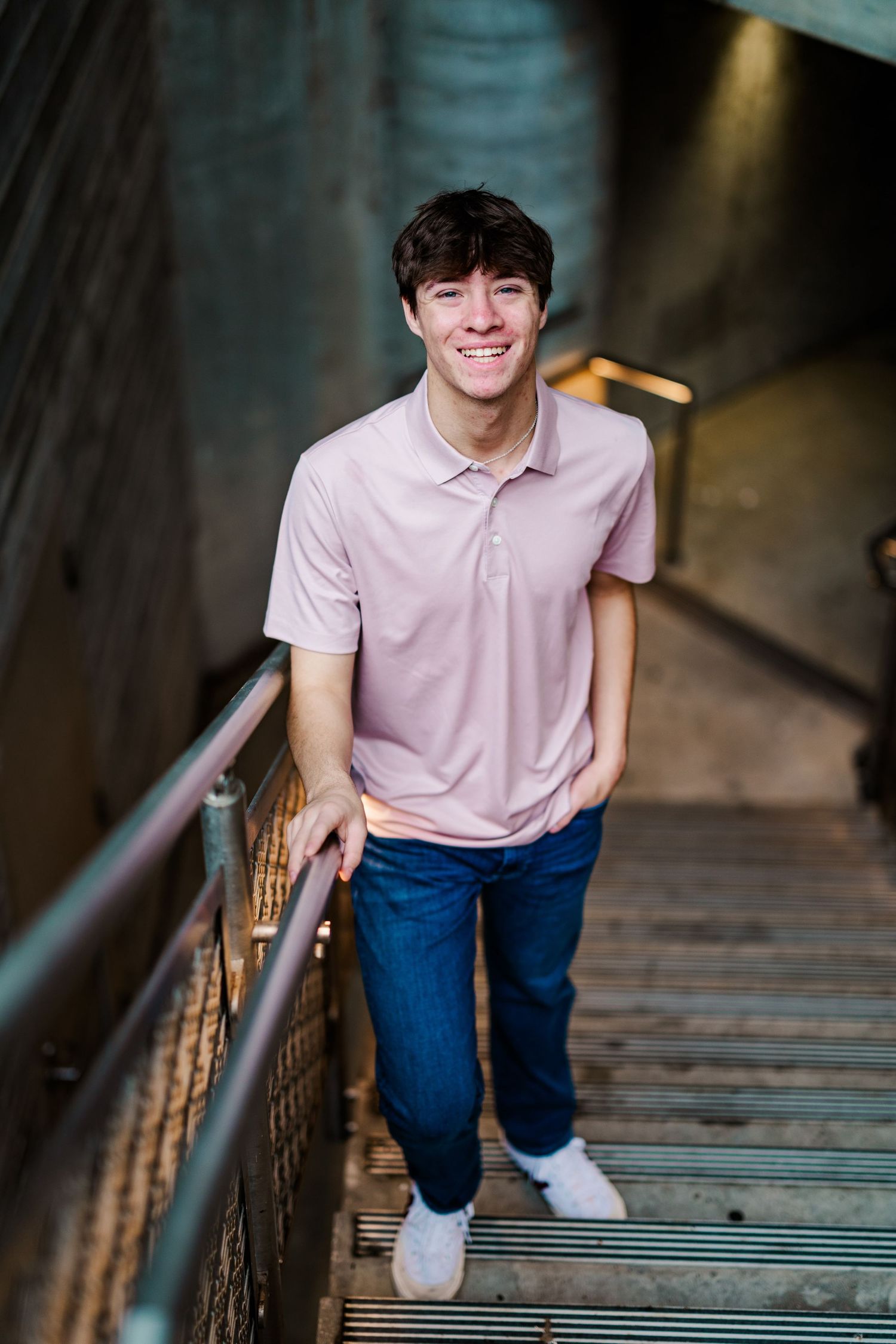 Smiling young man in pink polo shirt standing on industrial stairs holding metal railing.