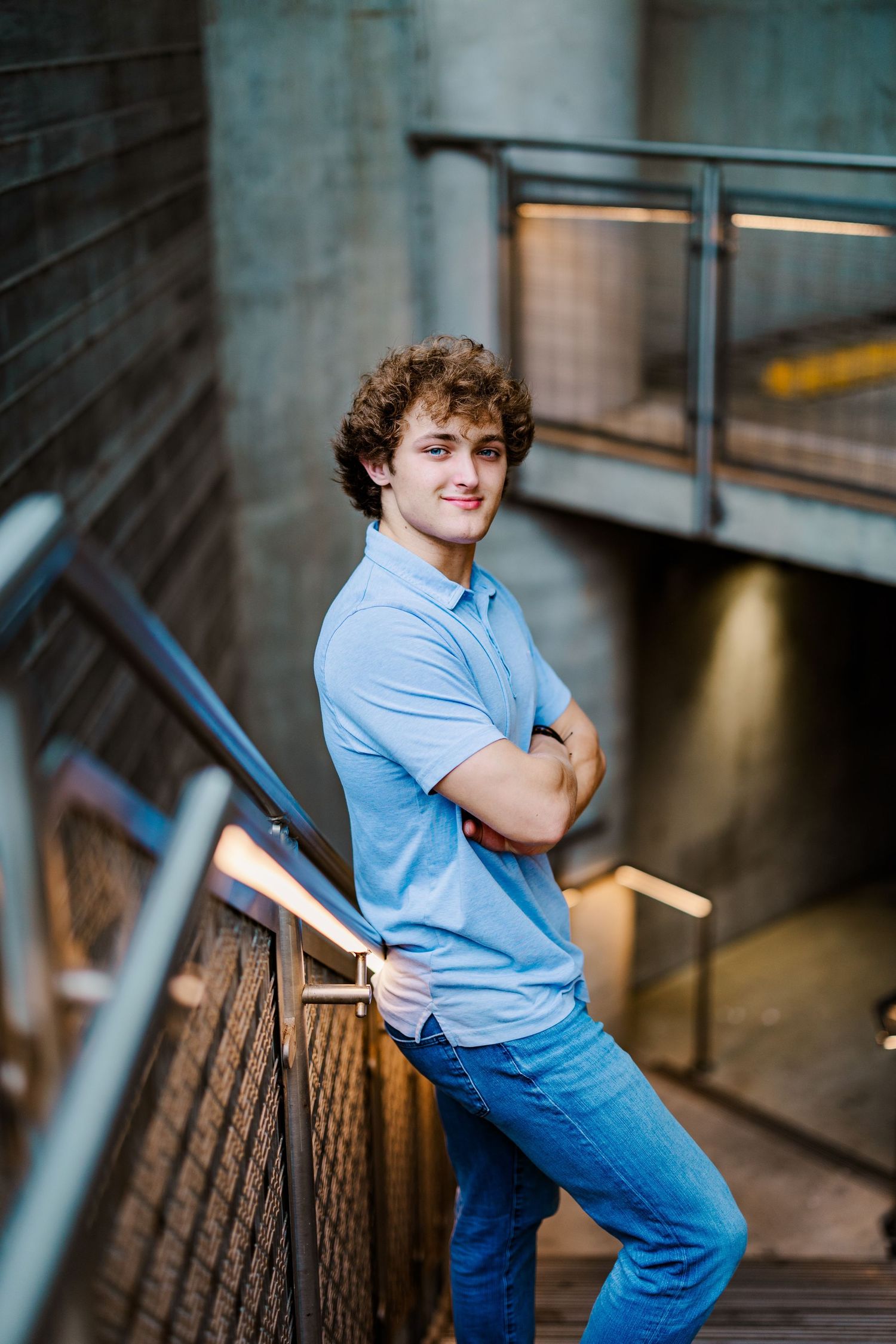 Young man with curly hair in blue polo shirt leans against metal railing on modern staircase.