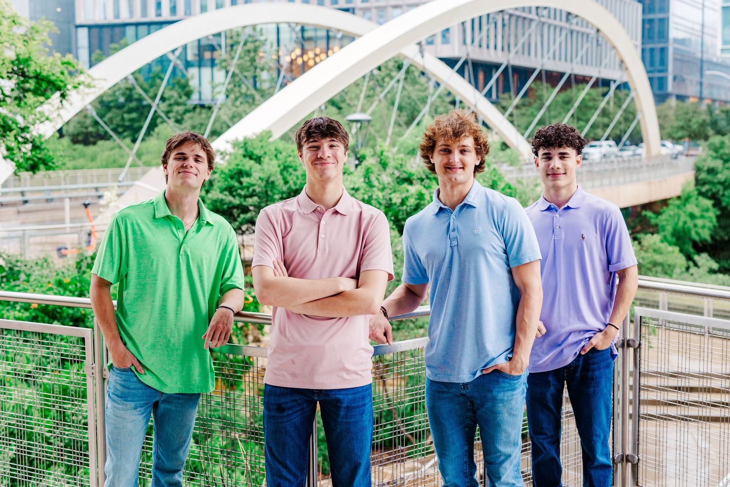 Four teenage boys in colorful polo shirts posing together on a bridge with city skyline in background.