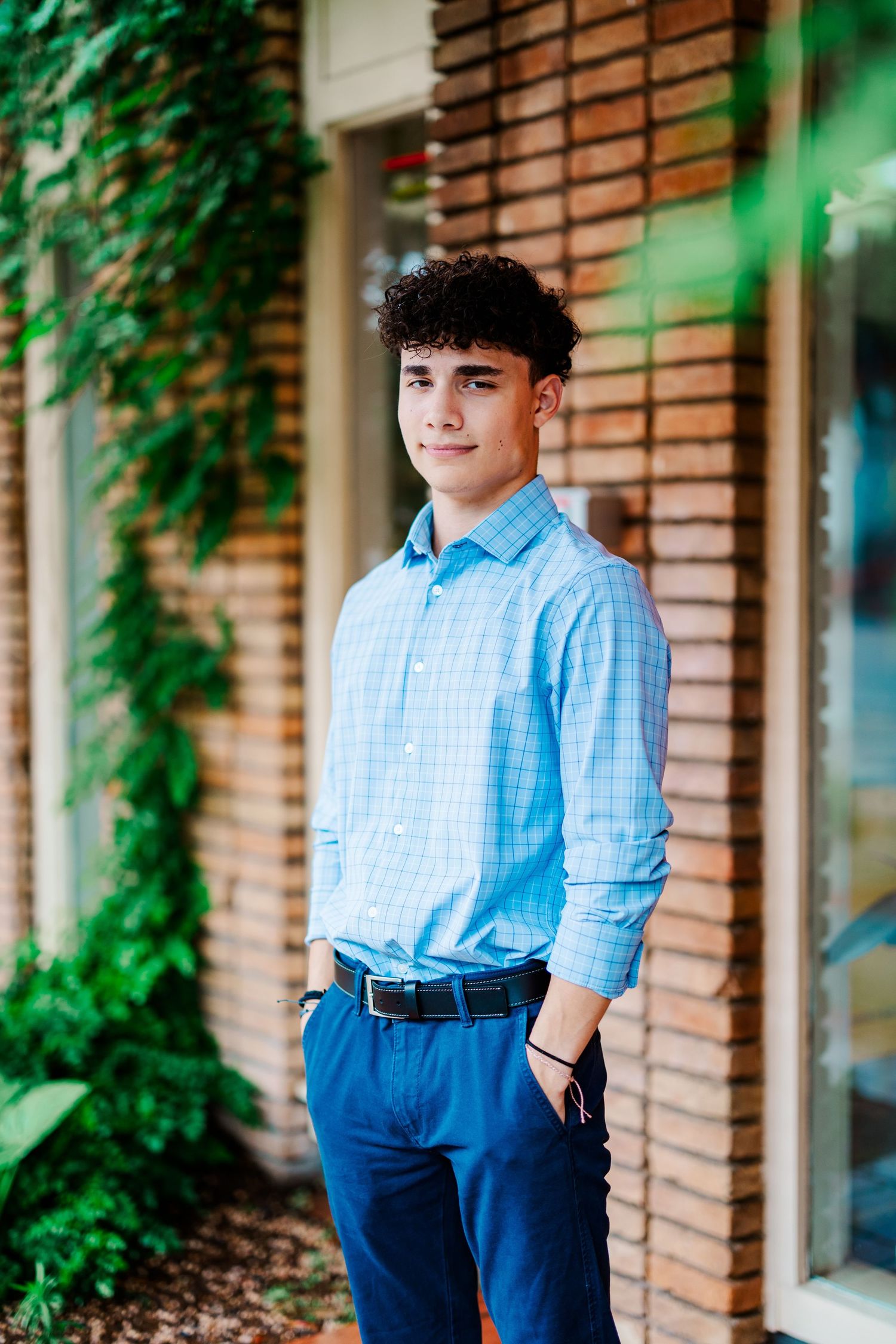 Teen boy in light blue plaid shirt and navy pants standing by ivy-covered brick wall.