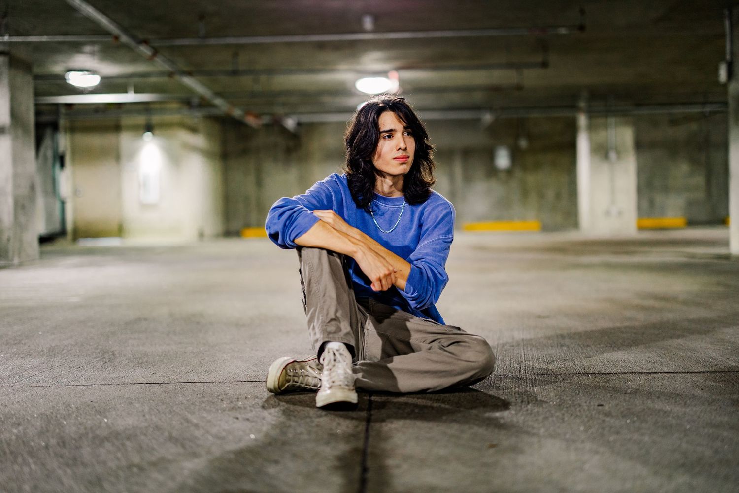 Young woman in blue shirt and gray pants sitting cross-legged on a parking garage floor.