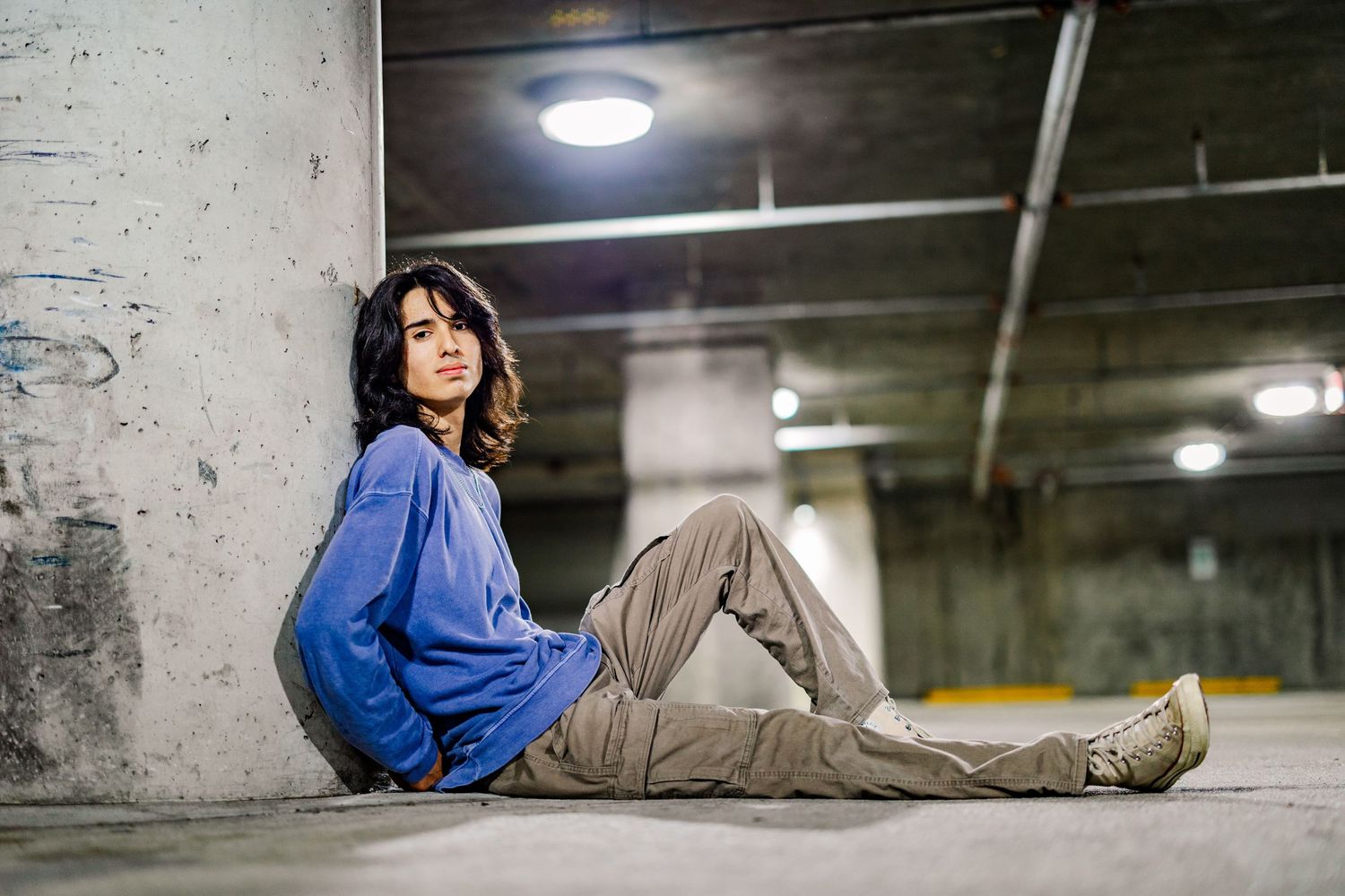 Young woman in blue hoodie and khaki pants sitting against concrete pillar in urban parking garage.