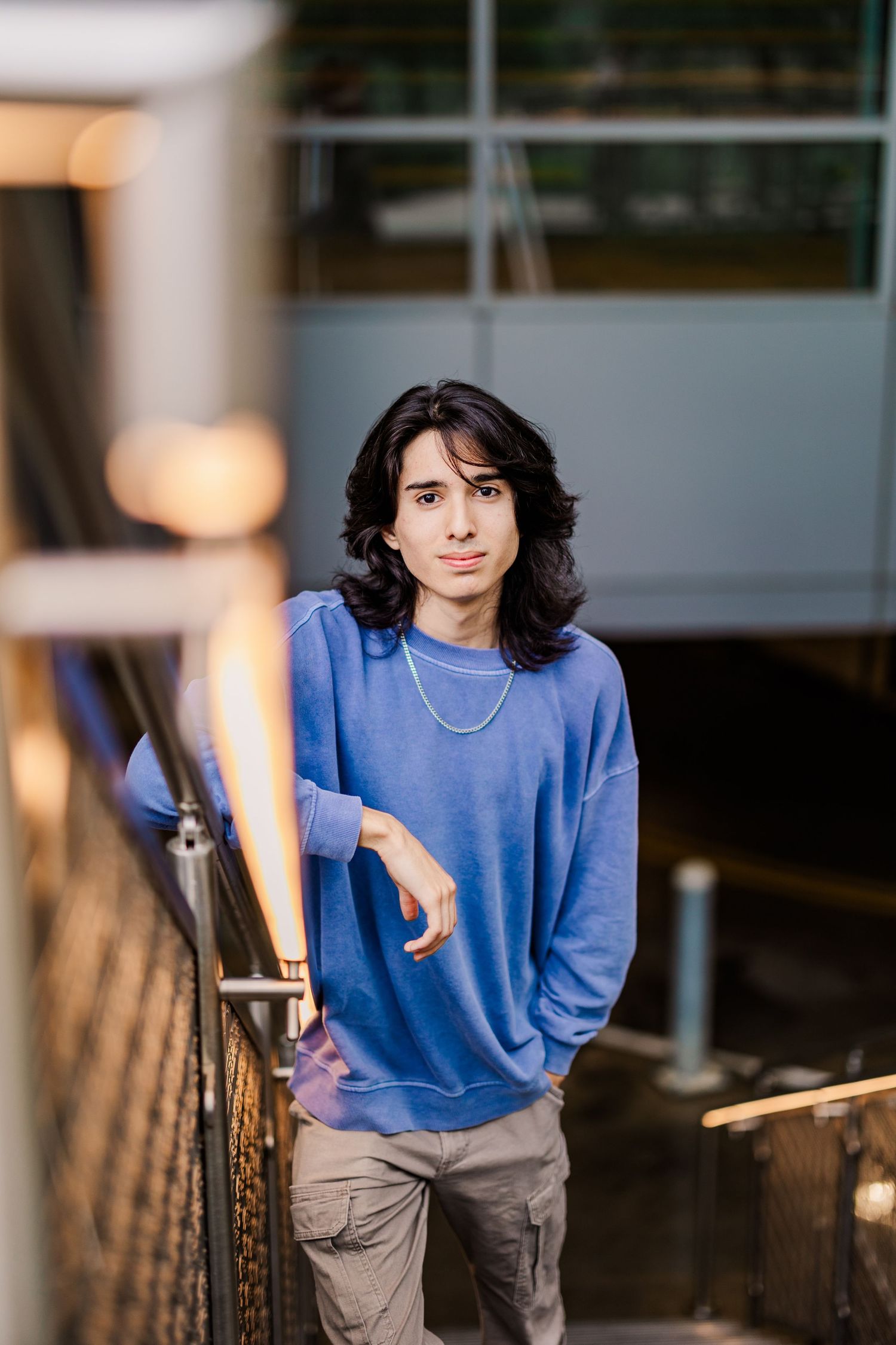 Young man with long dark hair in blue sweatshirt leaning on railing in urban setting.