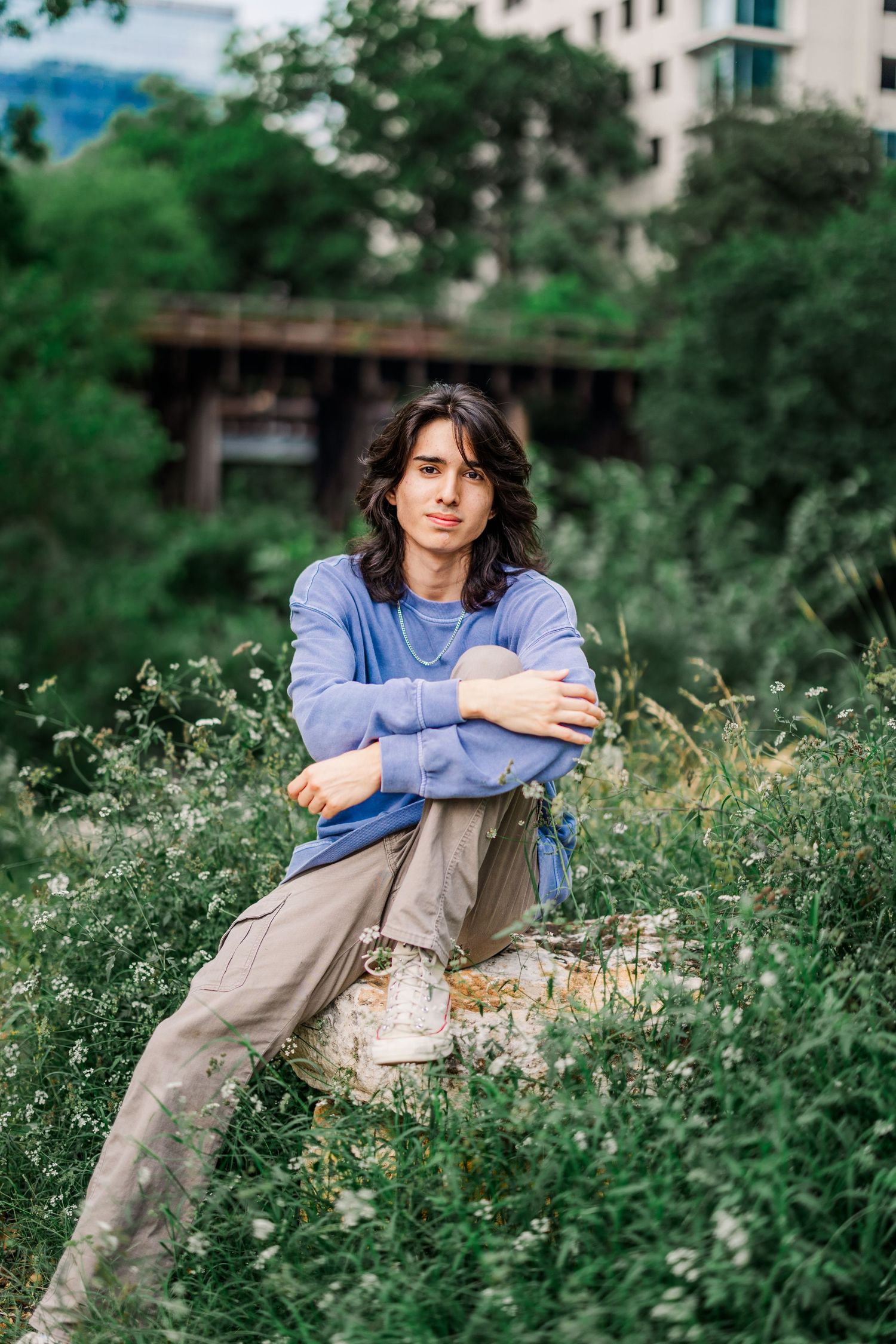 Young man with long dark hair in blue sweater sits cross-armed on stone wall among wildflowers.