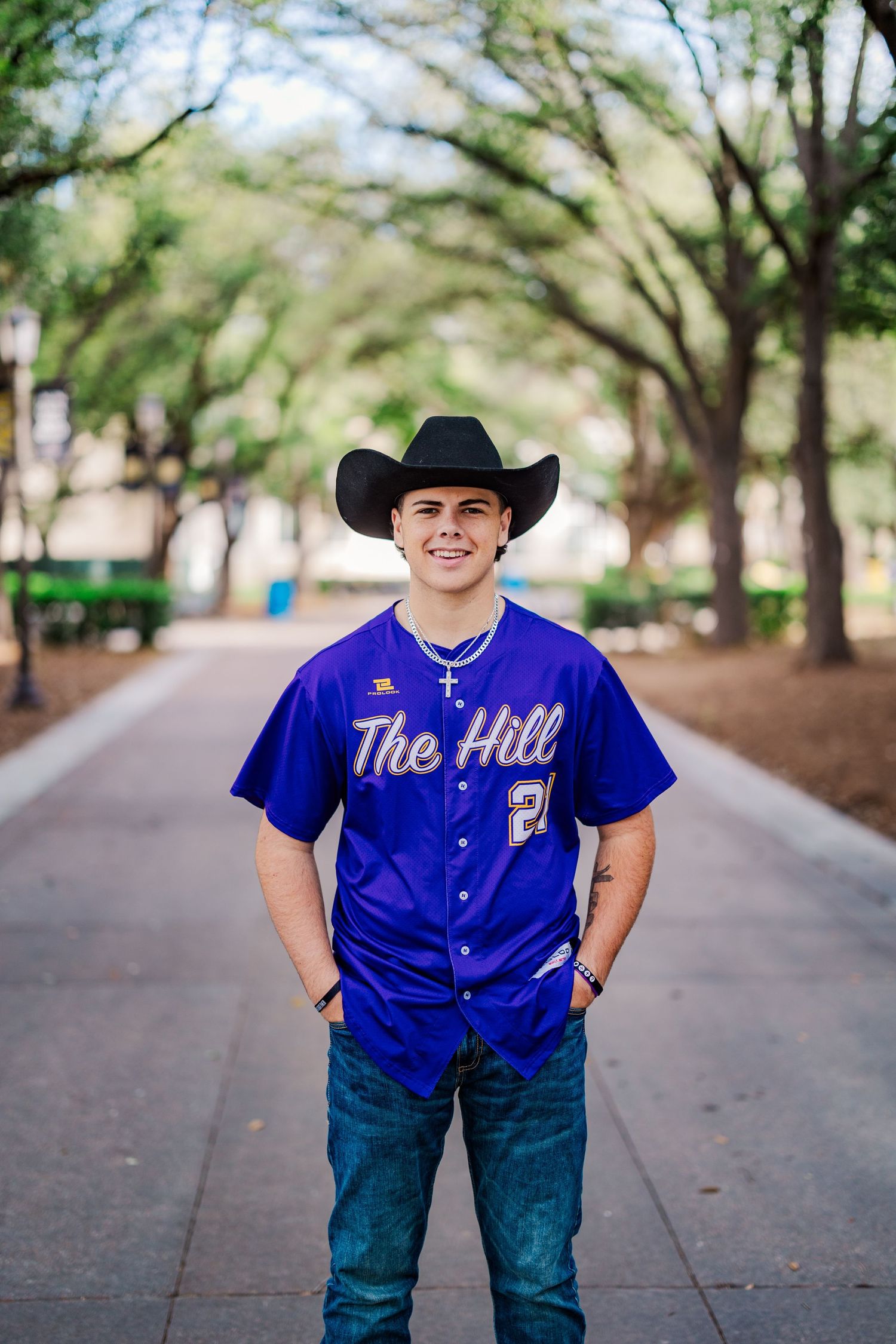Young man in purple The Hill baseball jersey and black cowboy hat on tree-lined path.