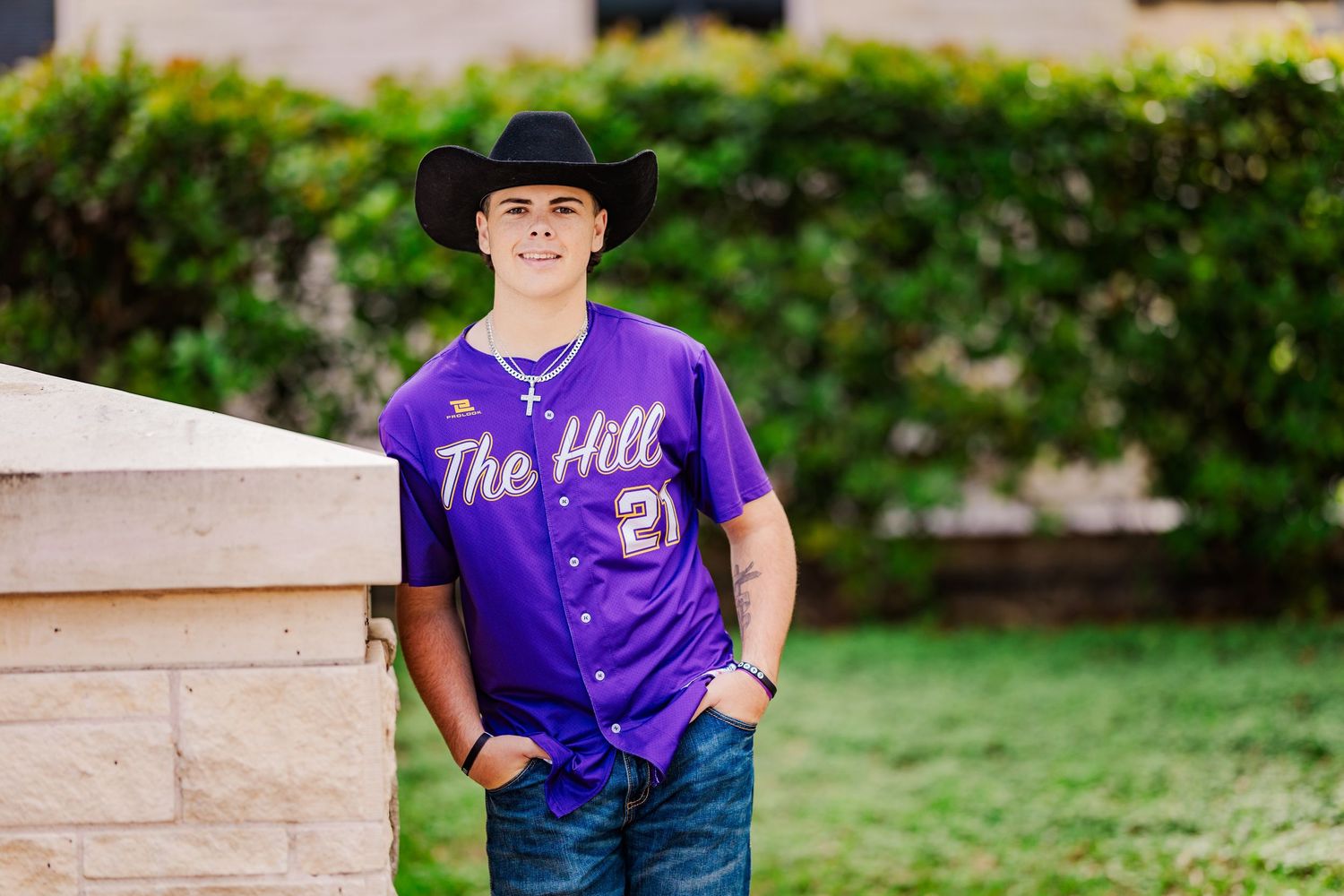 Smiling teen boy in purple baseball jersey and black cowboy hat posing outdoors.