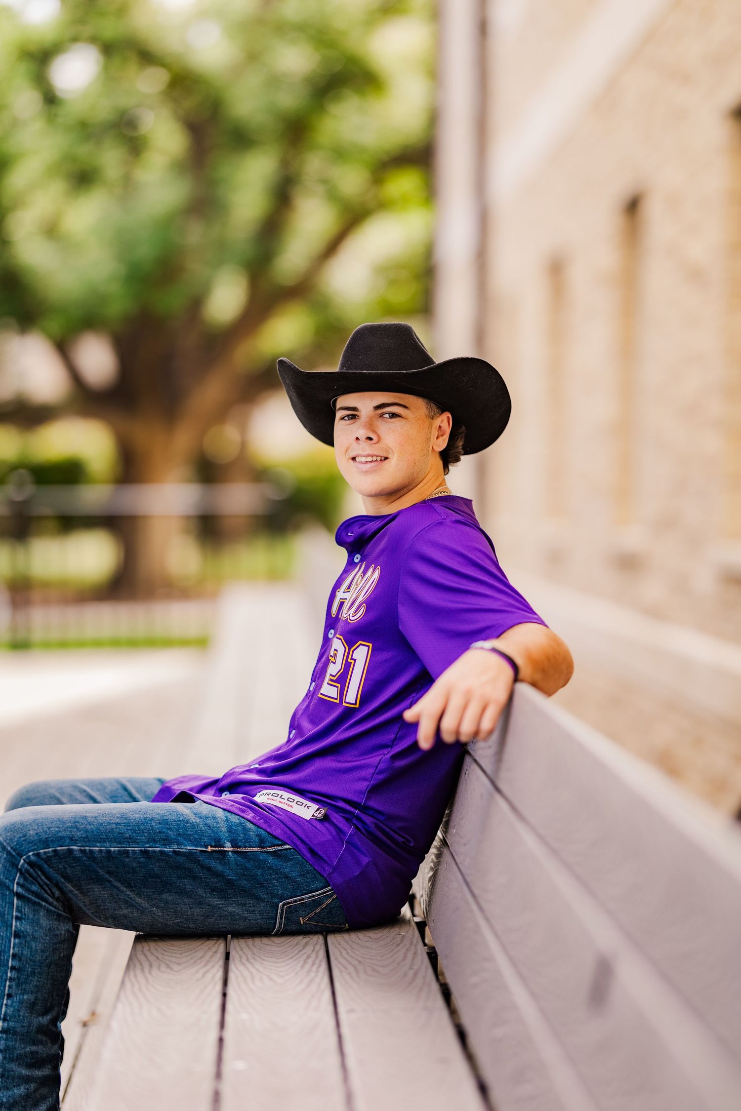 Teen boy in purple jersey and black cowboy hat sits relaxed on wooden bench smiling outdoors.