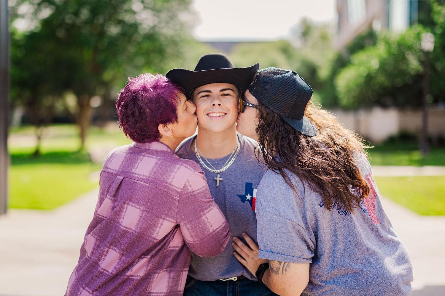 Three friends smile outdoors as two girls kiss a smiling teen wearing a black cowboy hat on the cheeks.