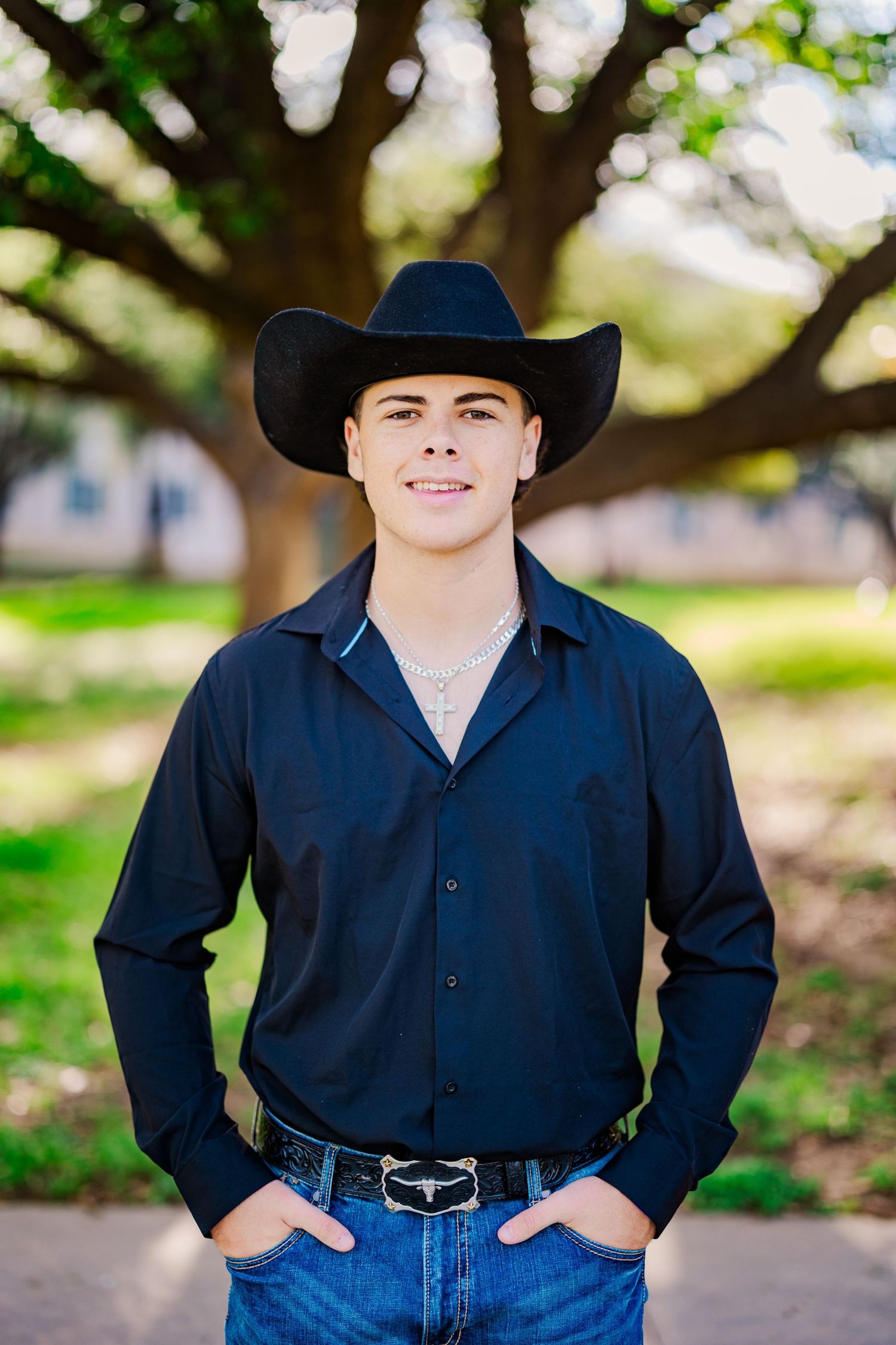Young man in black shirt and cowboy hat smiling outdoors under large oak tree.