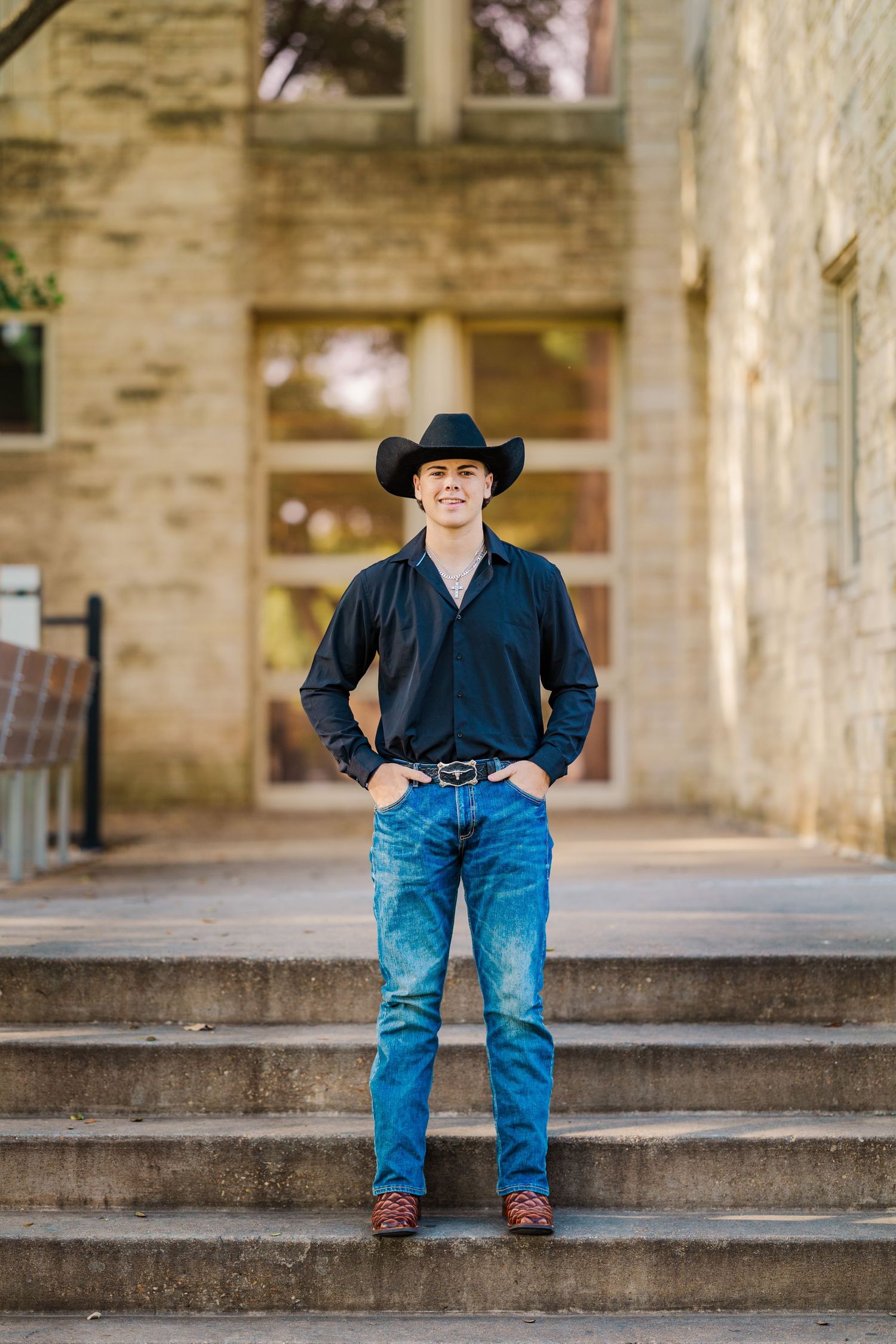 Young man in black shirt and cowboy hat standing on stone steps of historic building.