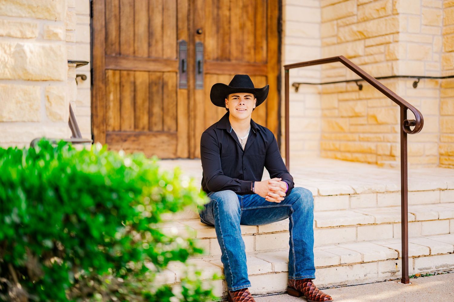 Smiling young man in cowboy hat sitting on stone steps outside rustic building entrance.