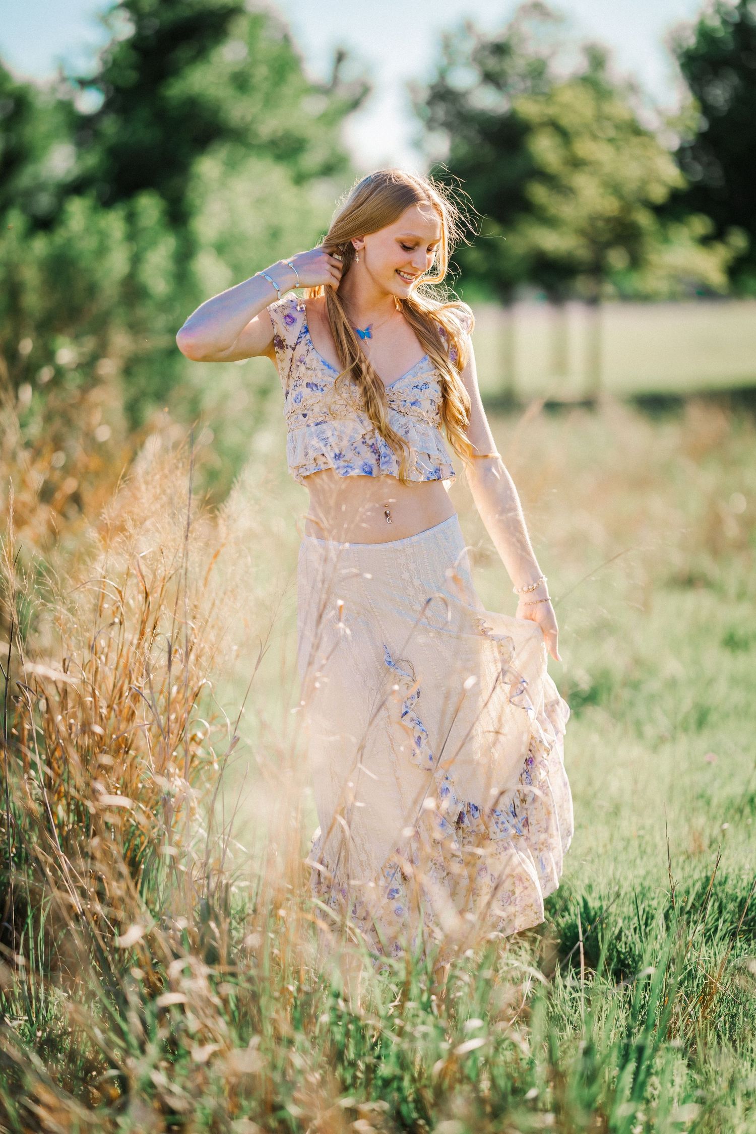 Blonde teenage girl in floral crop top and skirt standing in a sunlit golden grass field.