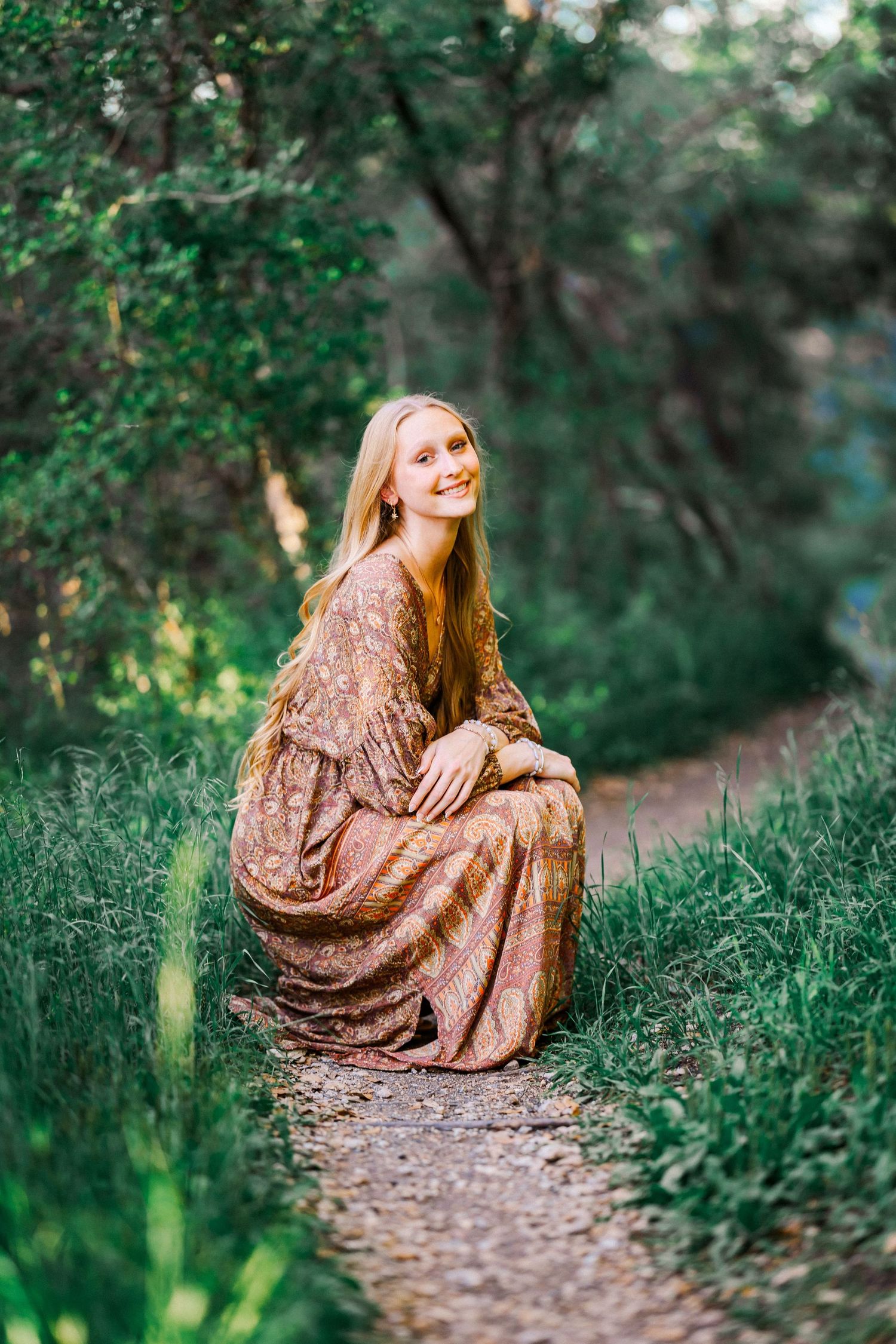 Young woman with long red hair sitting on a forest path in a floral bohemian dress, smiling outdoors.