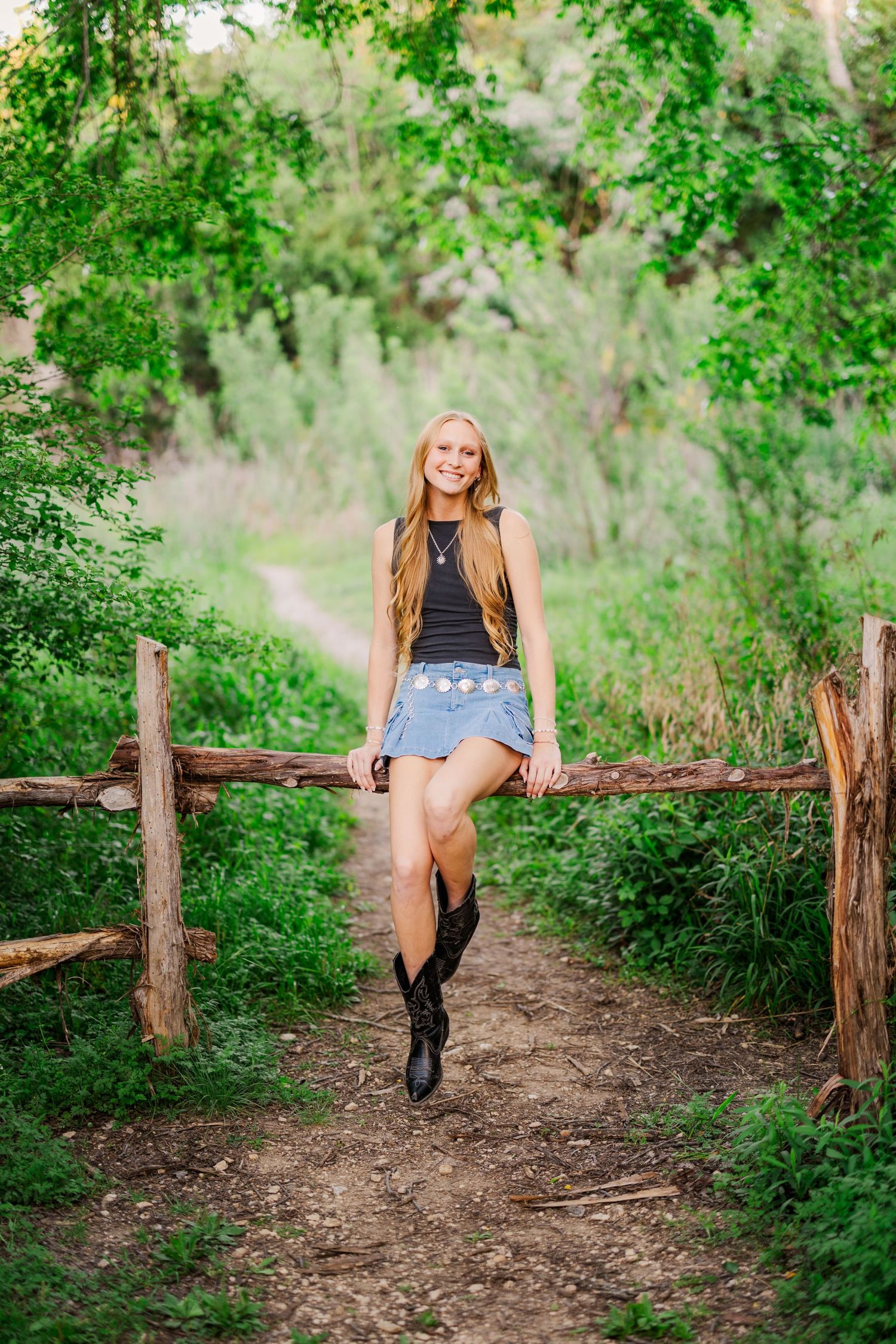 Smiling teenage girl in denim skirt and cowboy boots leaning on rustic wooden fence in lush green outdoor setting.