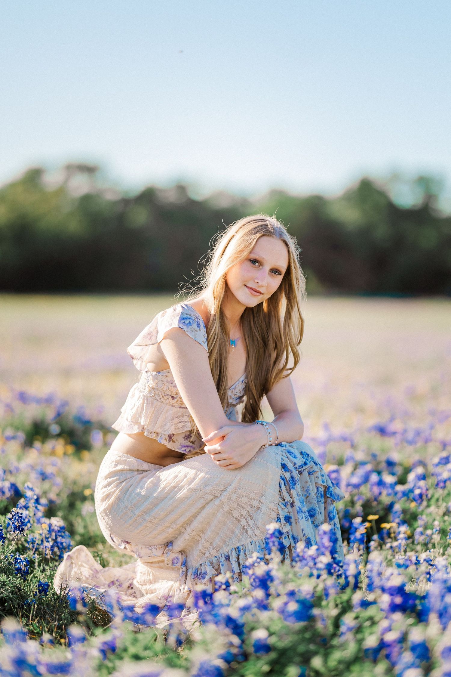 Blonde teenage girl crouches in vibrant bluebonnet wildflower field under clear blue sky.