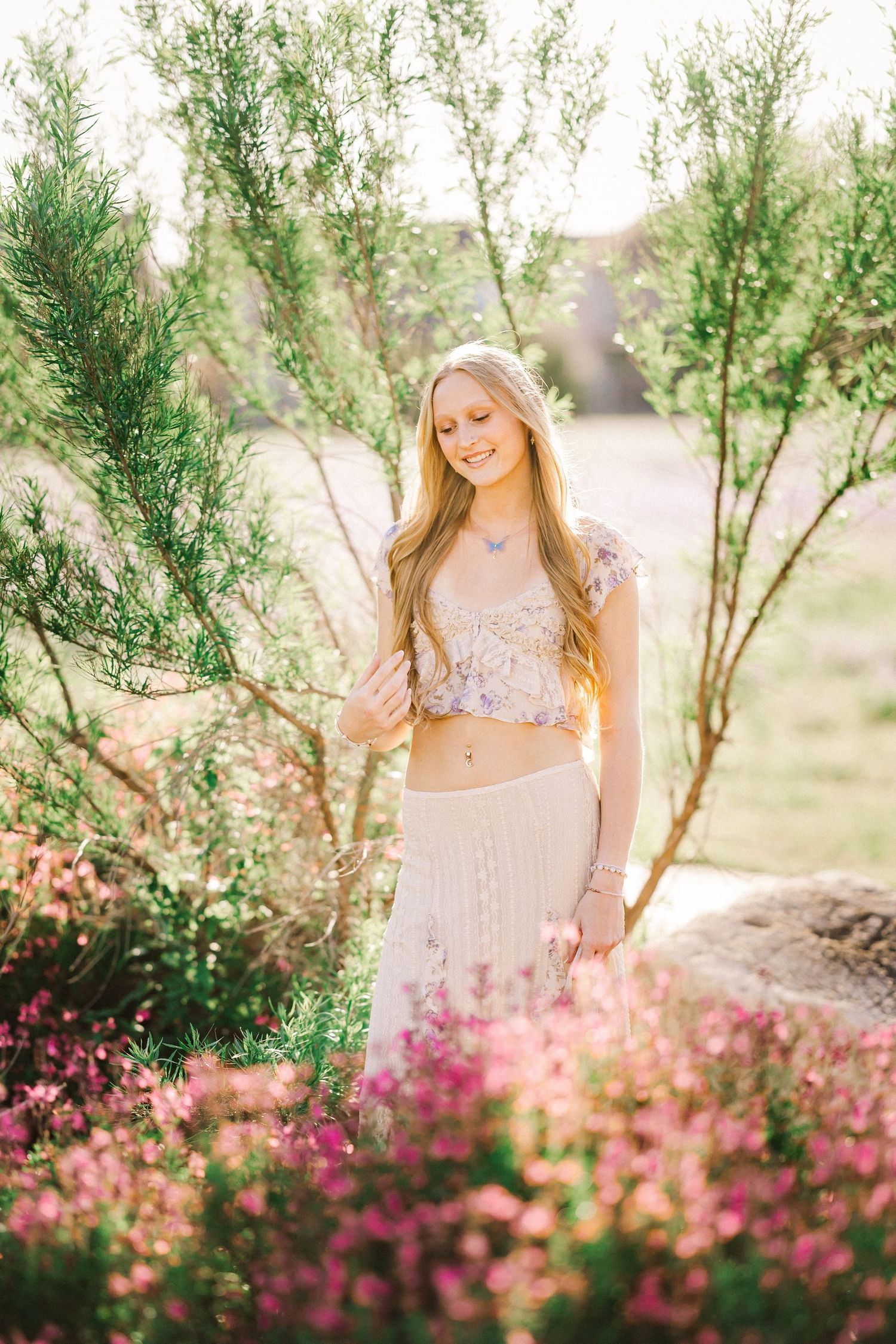 Smiling blonde girl in floral crop top and white skirt stands among pink flowers and green shrubs.