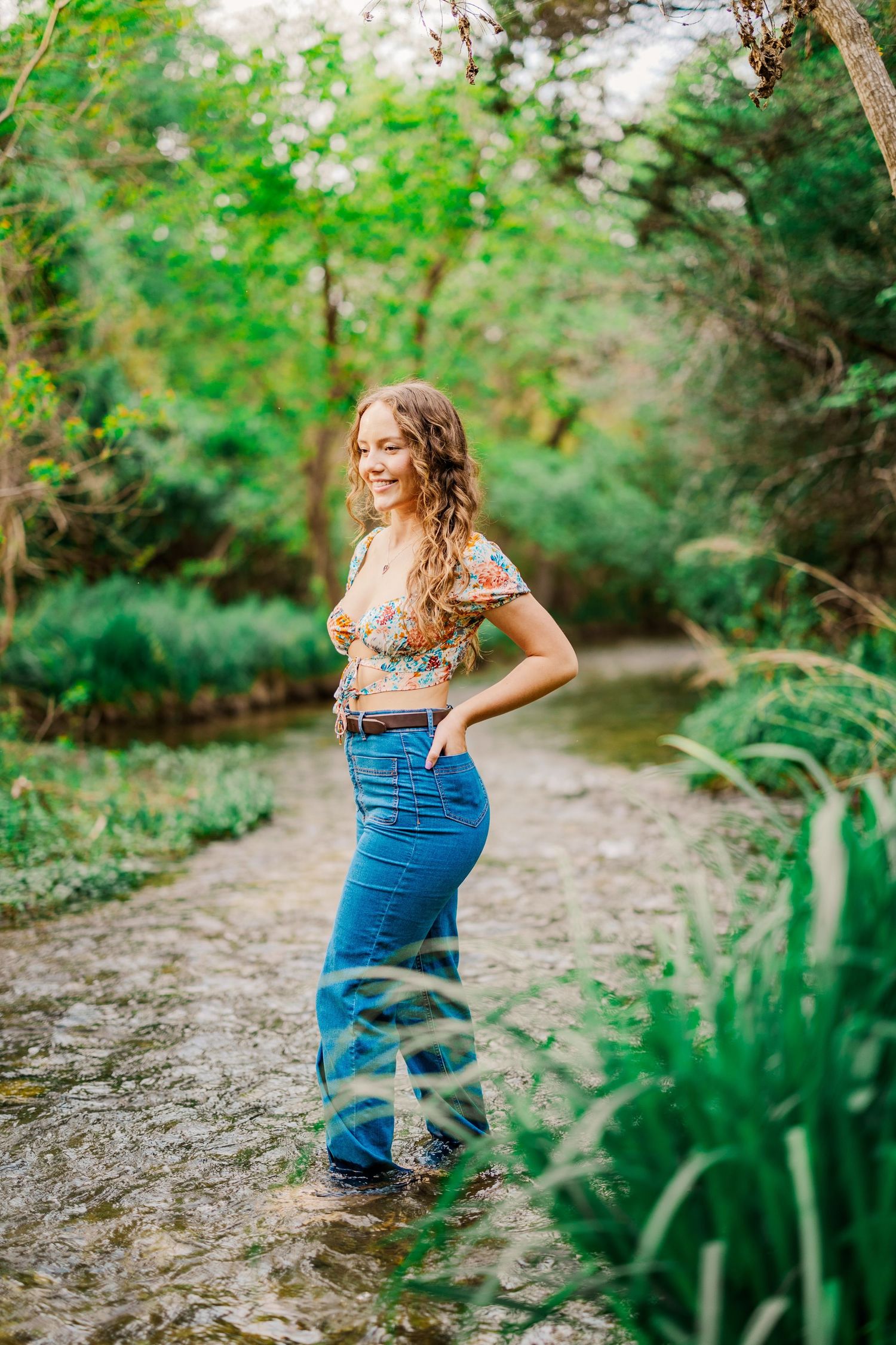 Young woman in floral top and wide-leg jeans stands in shallow creek surrounded by green trees.