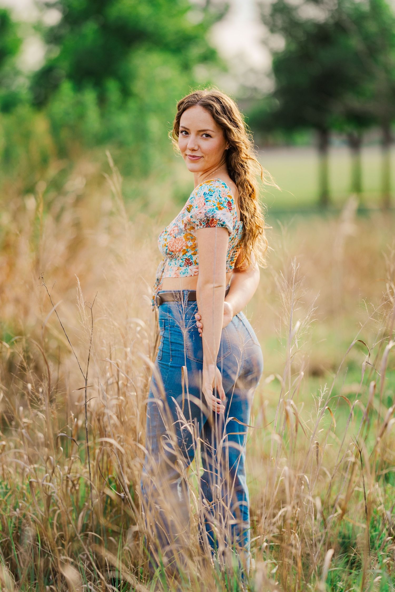 Young woman in floral crop top and jeans stands in tall golden grass field at golden hour.