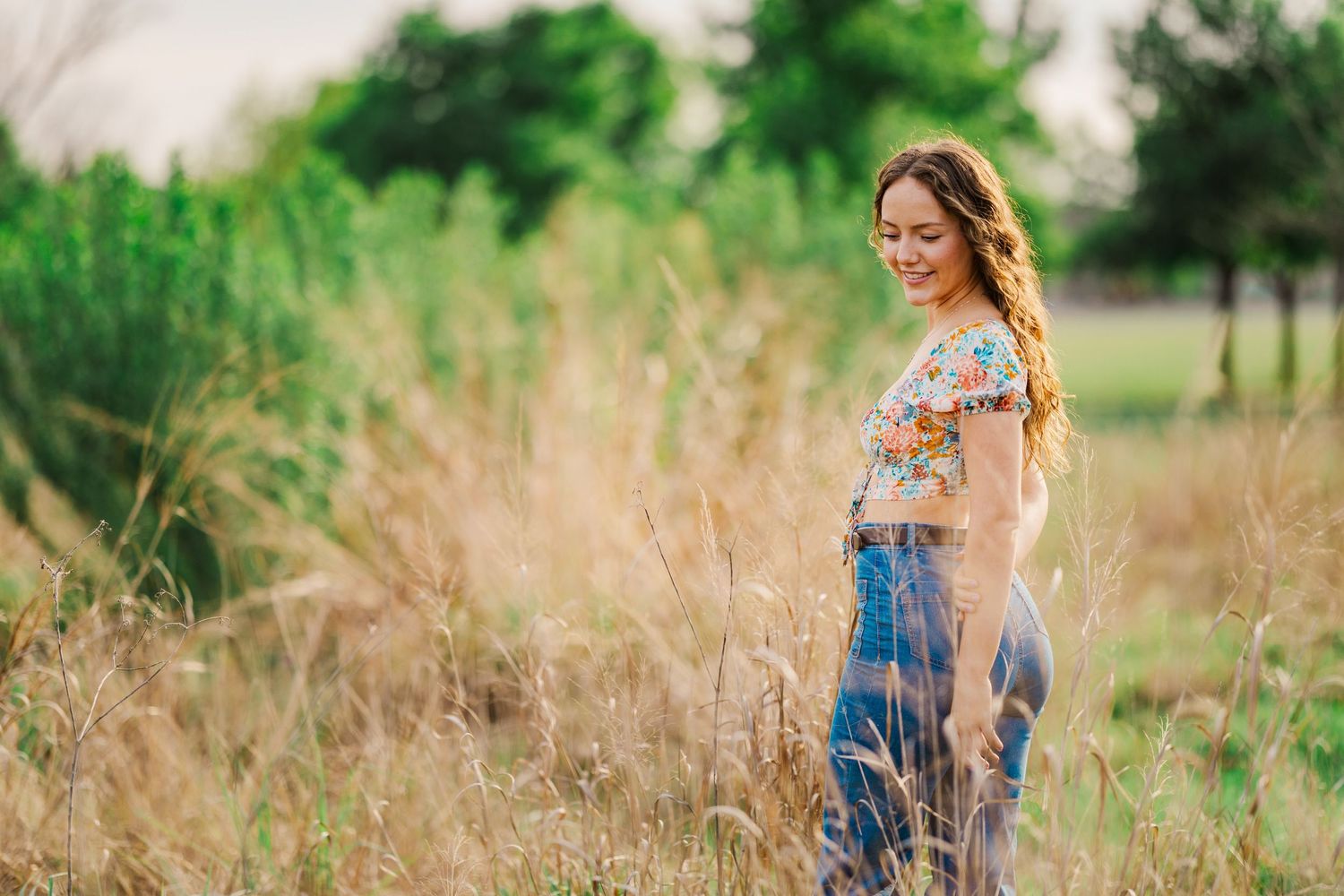 Young woman in floral top and jeans smiling in tall golden grass field at sunset.