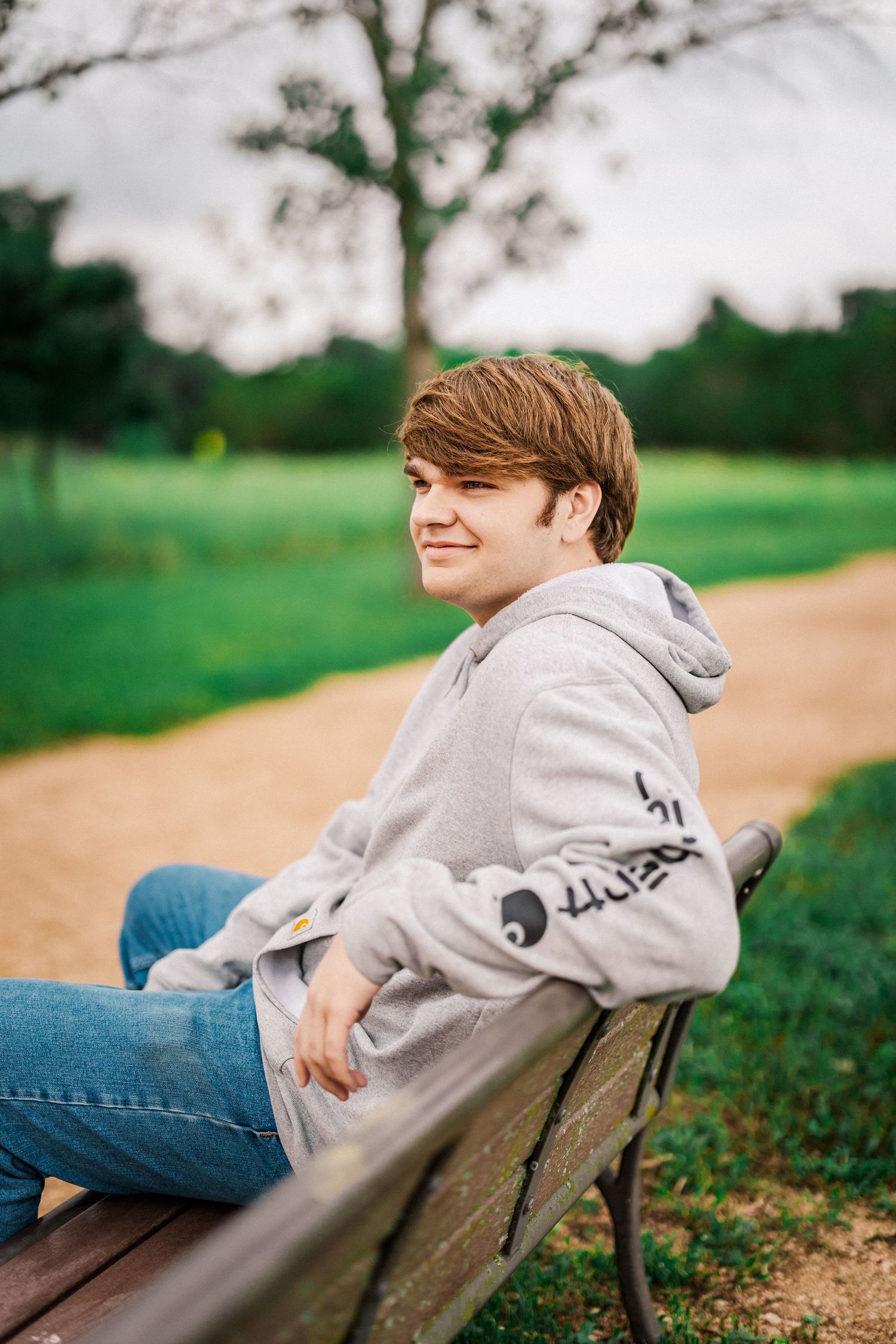Teenage boy in gray hoodie sitting on a park bench smiling with green grass and trees behind him.