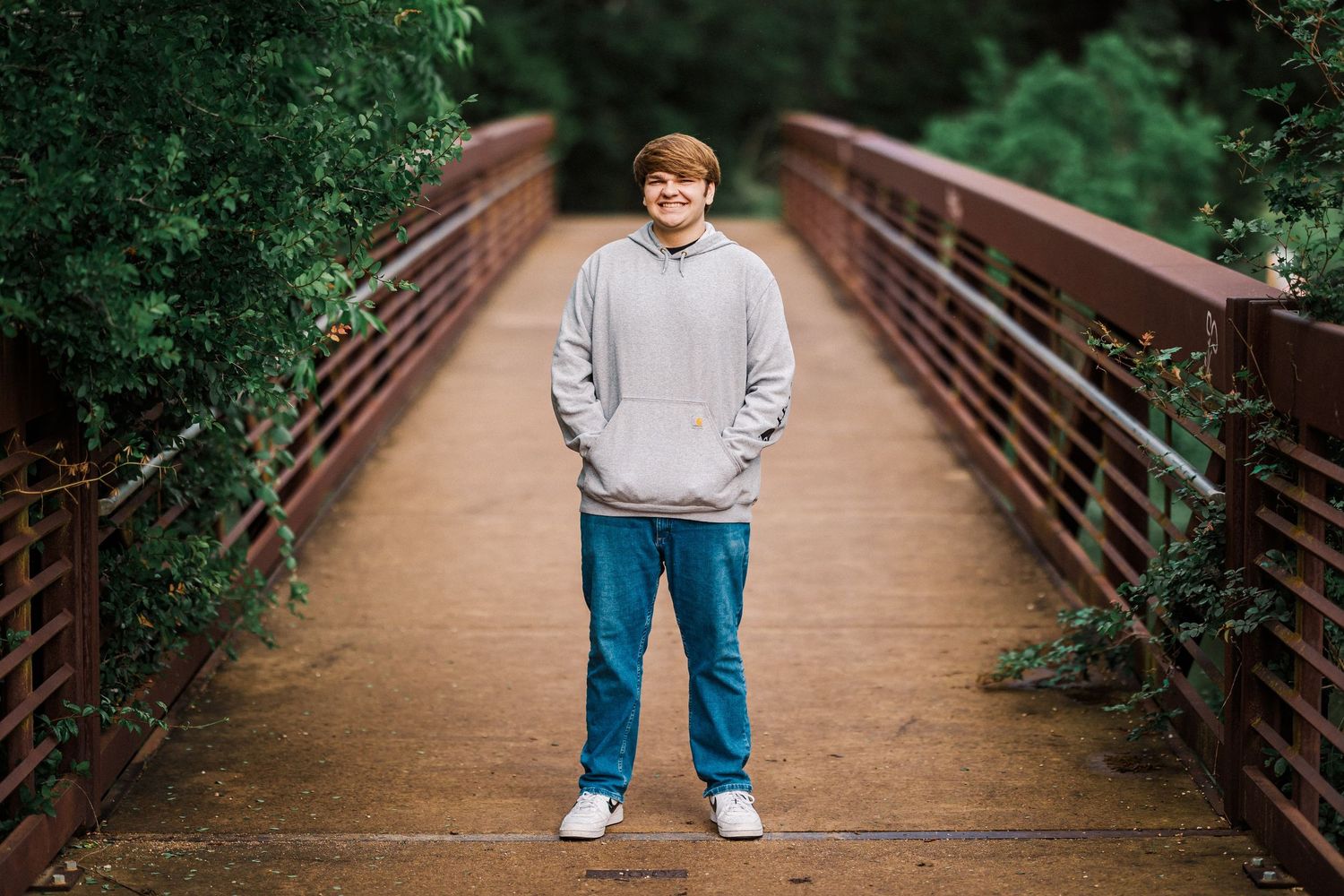 Smiling teen in a gray hoodie and jeans standing on a wooden bridge with greenery.