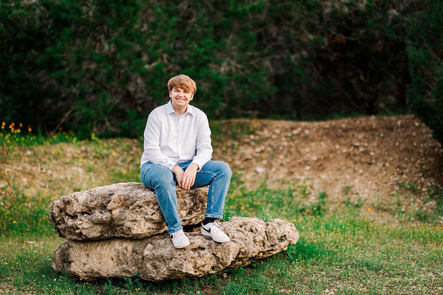 Red-haired teen boy in white shirt and jeans sitting on a large rock in a green outdoor setting.