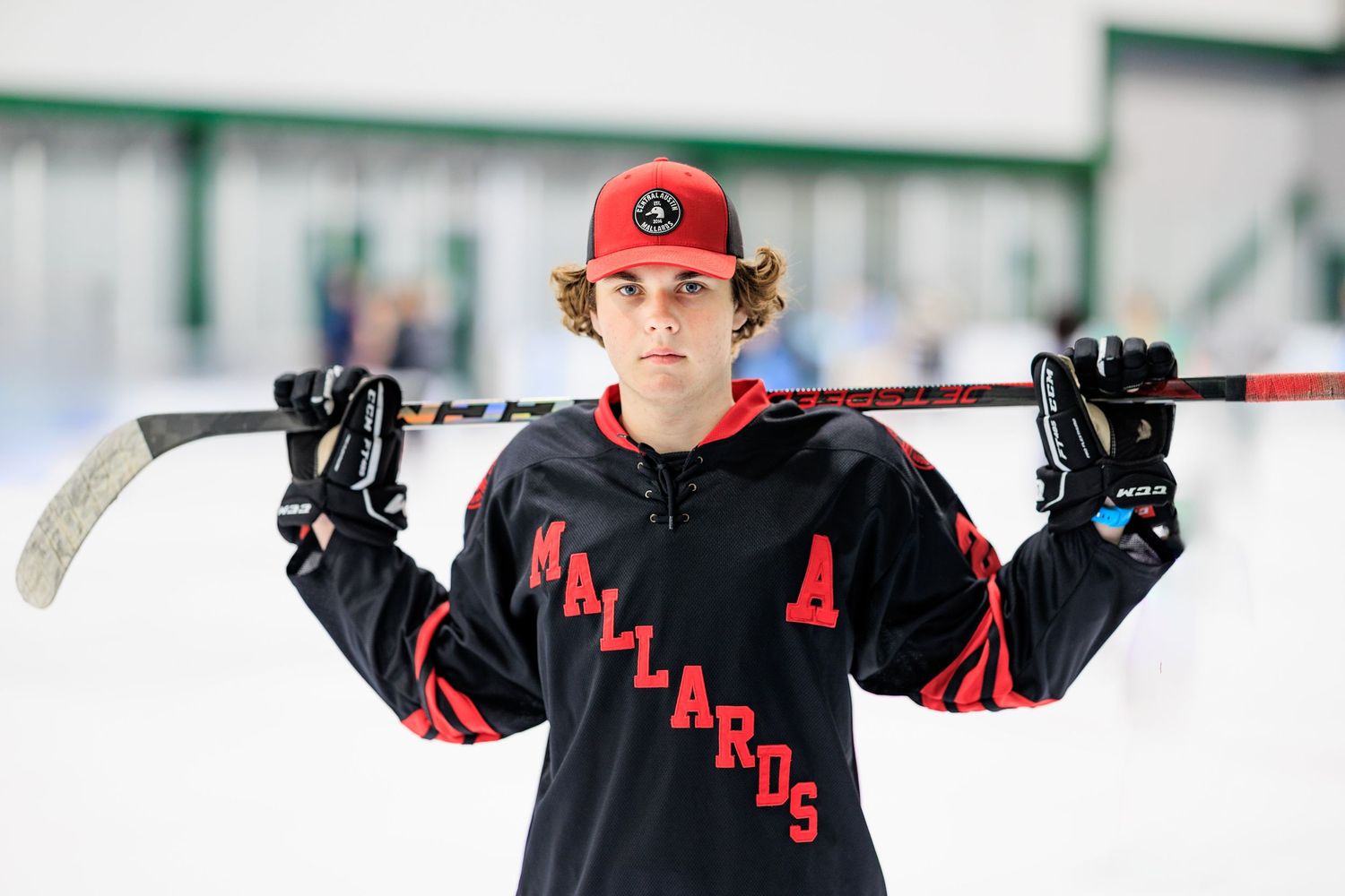 Teen hockey player in Mallards jersey poses on ice rink holding stick across shoulders with confident stance.