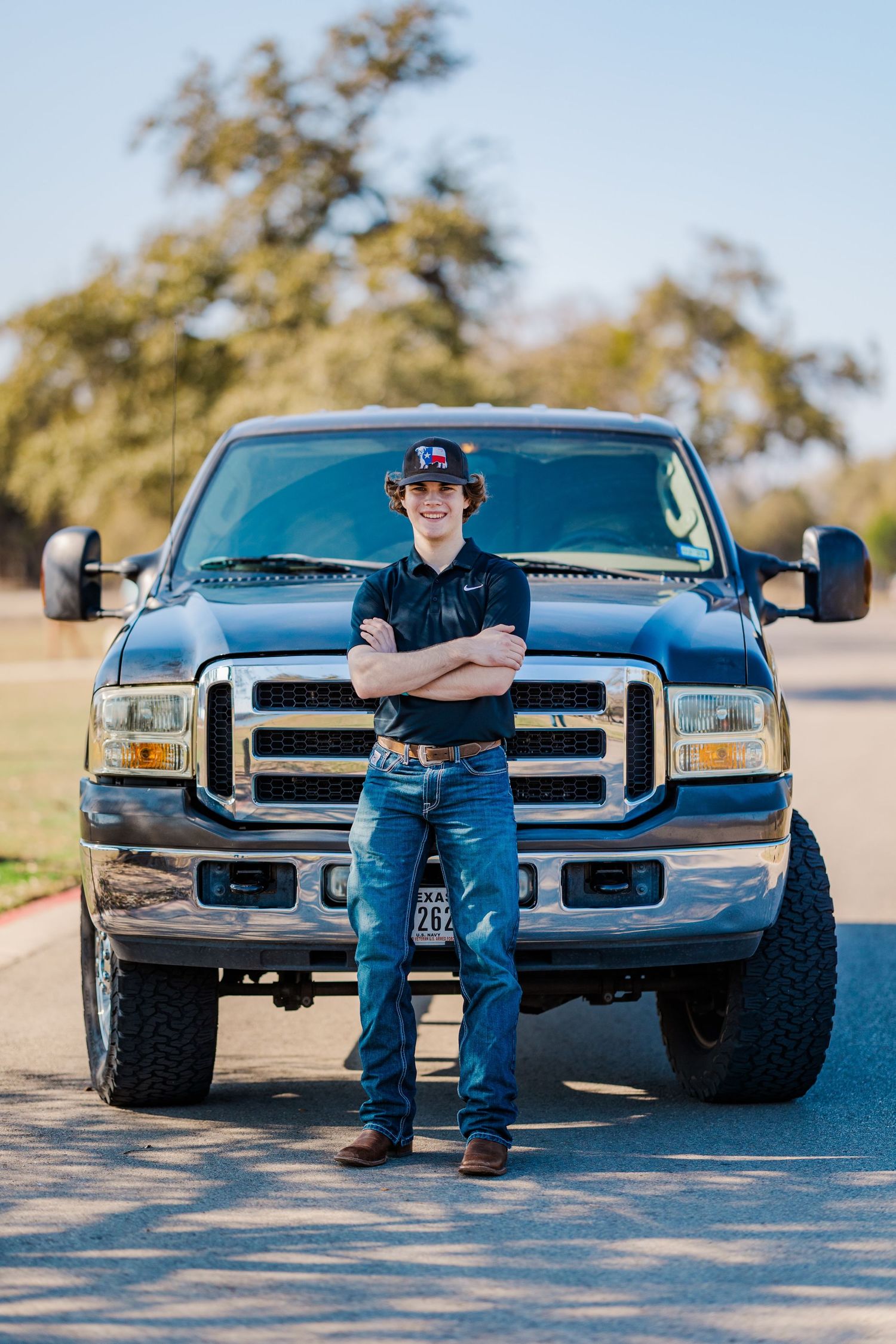 Teen boy in jeans and cap standing arms crossed before black Ford pickup truck.