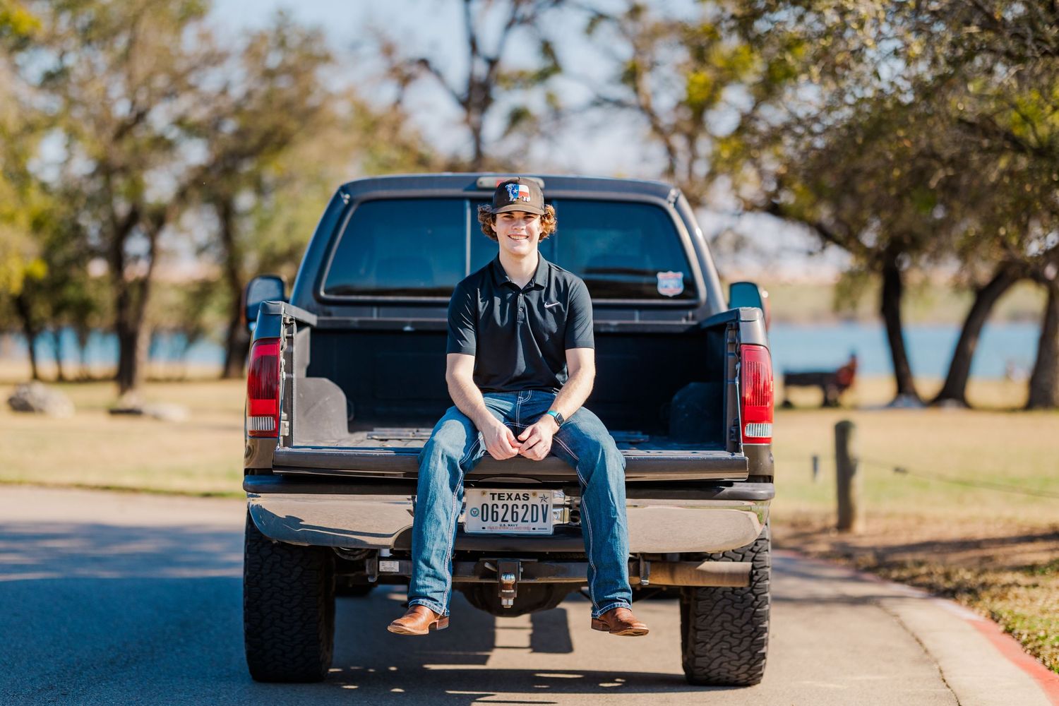 Young man in baseball cap sitting on truck tailgate parked by lakeside park.
