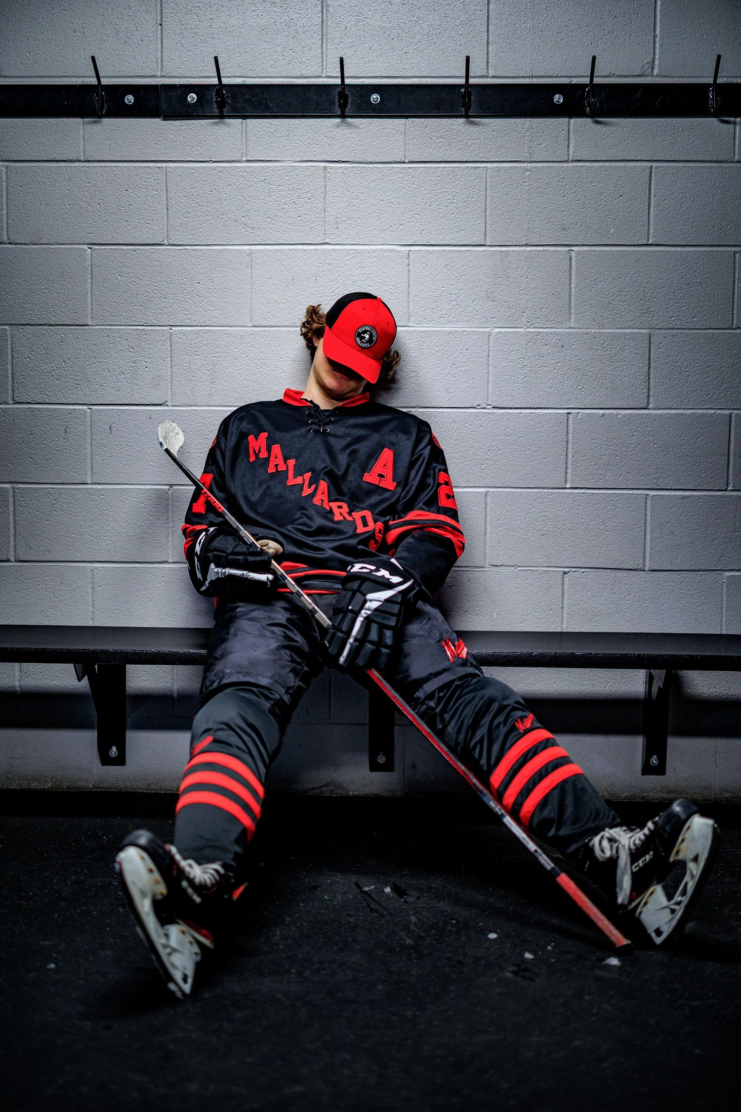 Hockey player in black and red uniform sitting in locker room with stick and skates.