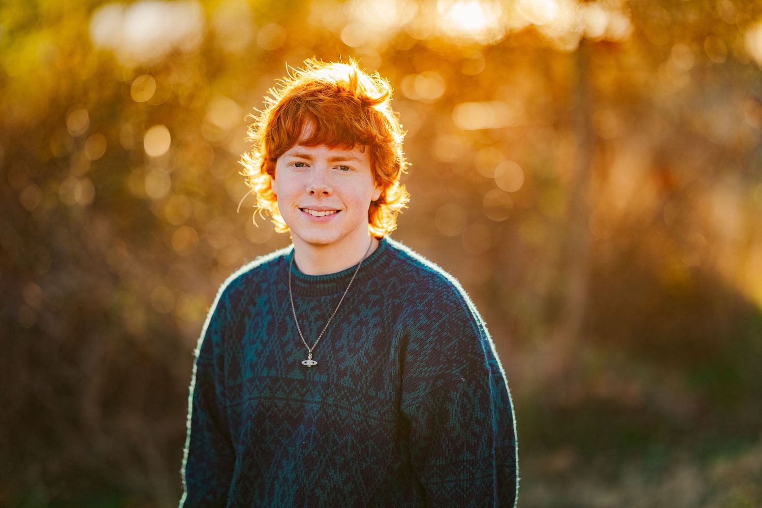 Smiling teenage boy with red curly hair wearing dark sweater and necklace in golden sunset light.