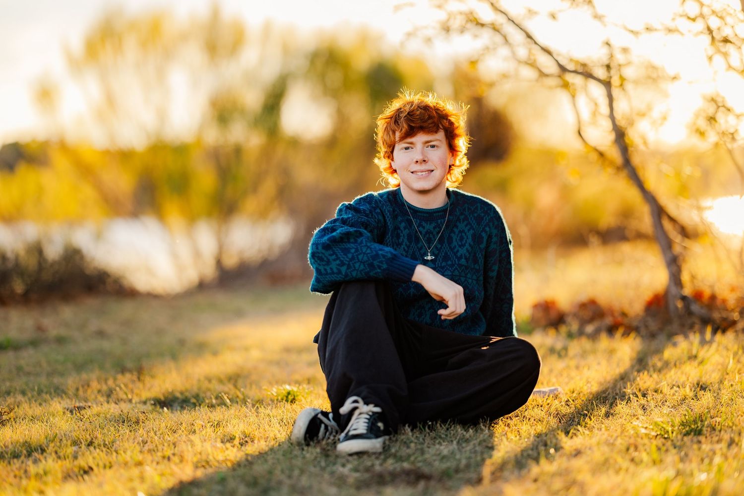 Red-haired teen boy sitting cross-legged on grass during golden hour sunset outdoors.