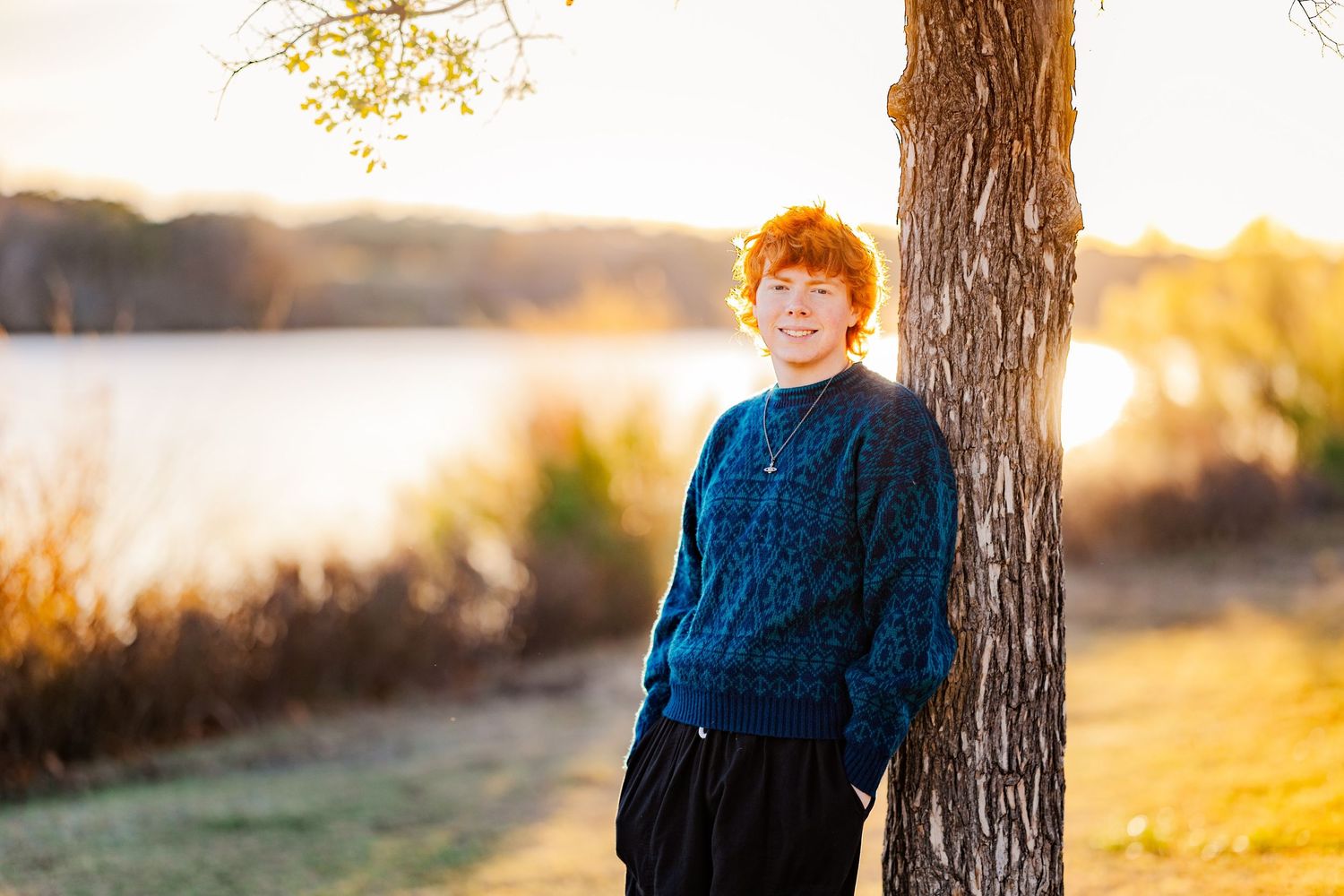 Smiling teen with red curly hair leans against tree in blue sweater near sunlit lake in autumn.