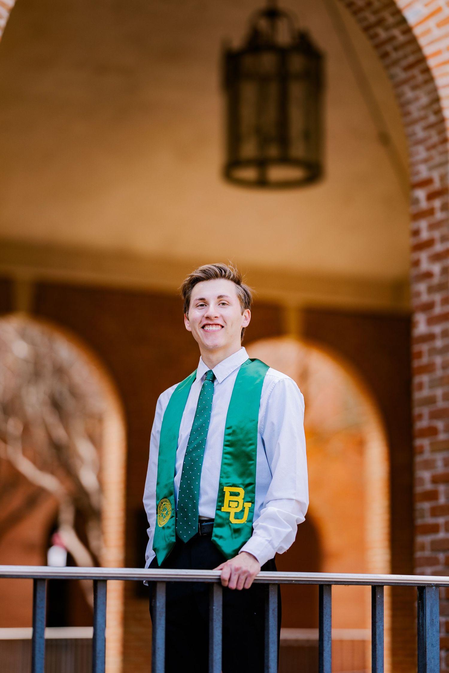 Male graduate wearing Baylor University green stole and white dress shirt posing on campus balcony.