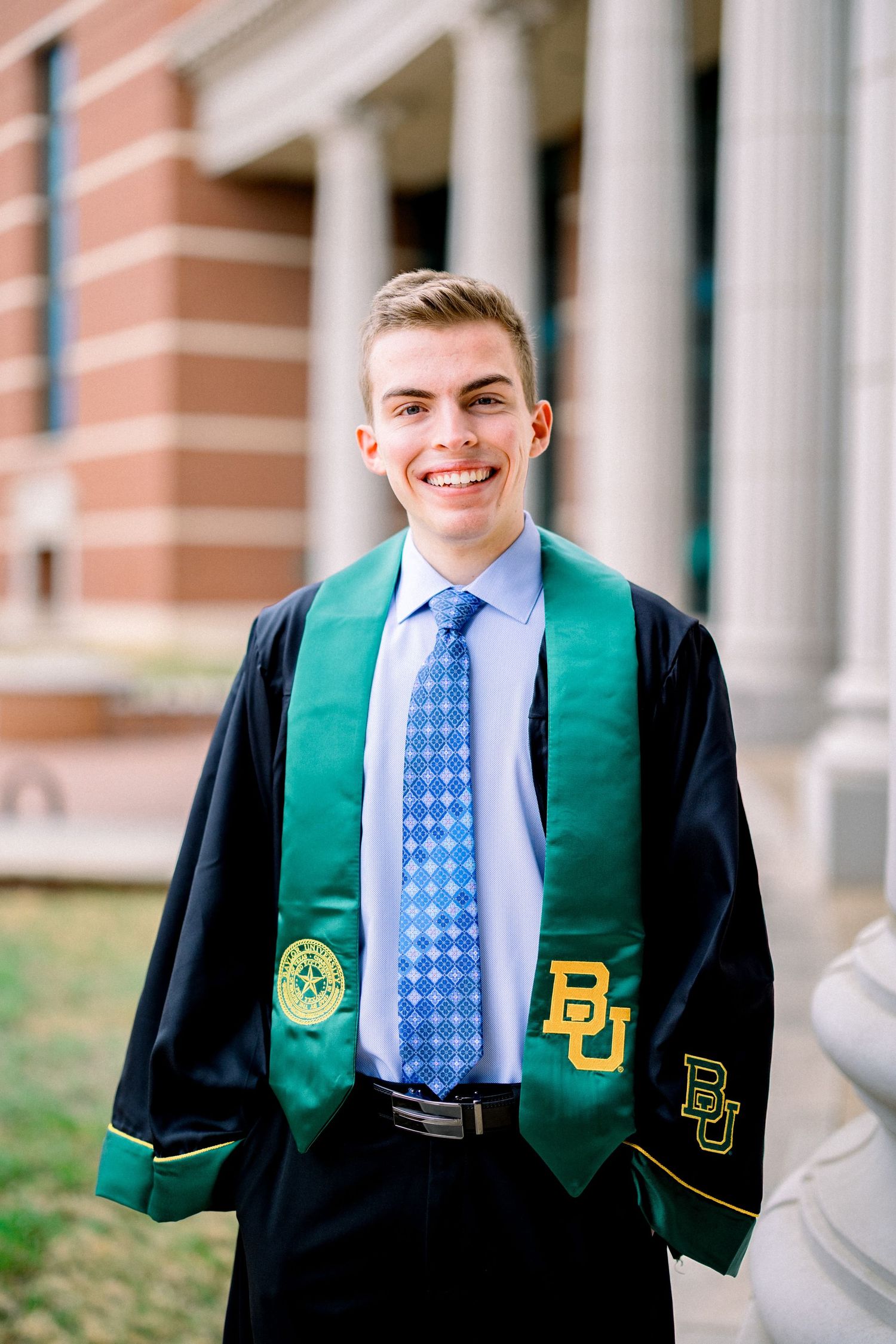 Smiling Baylor University graduate in black graduation gown with green BU stole poses outdoors on campus.