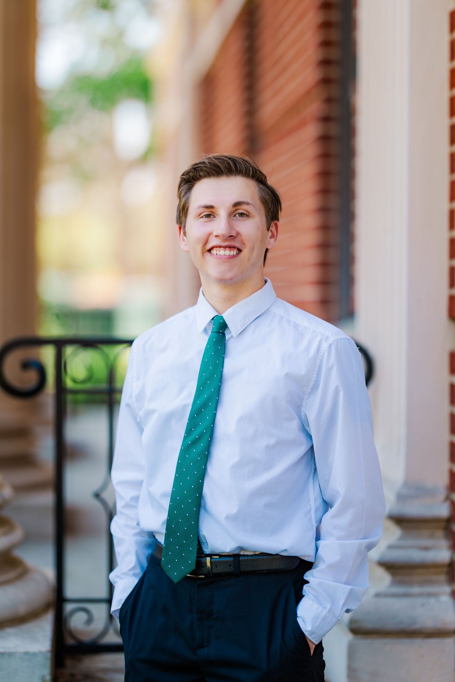 Smiling young man in white dress shirt and teal tie posing between columns for senior portrait.