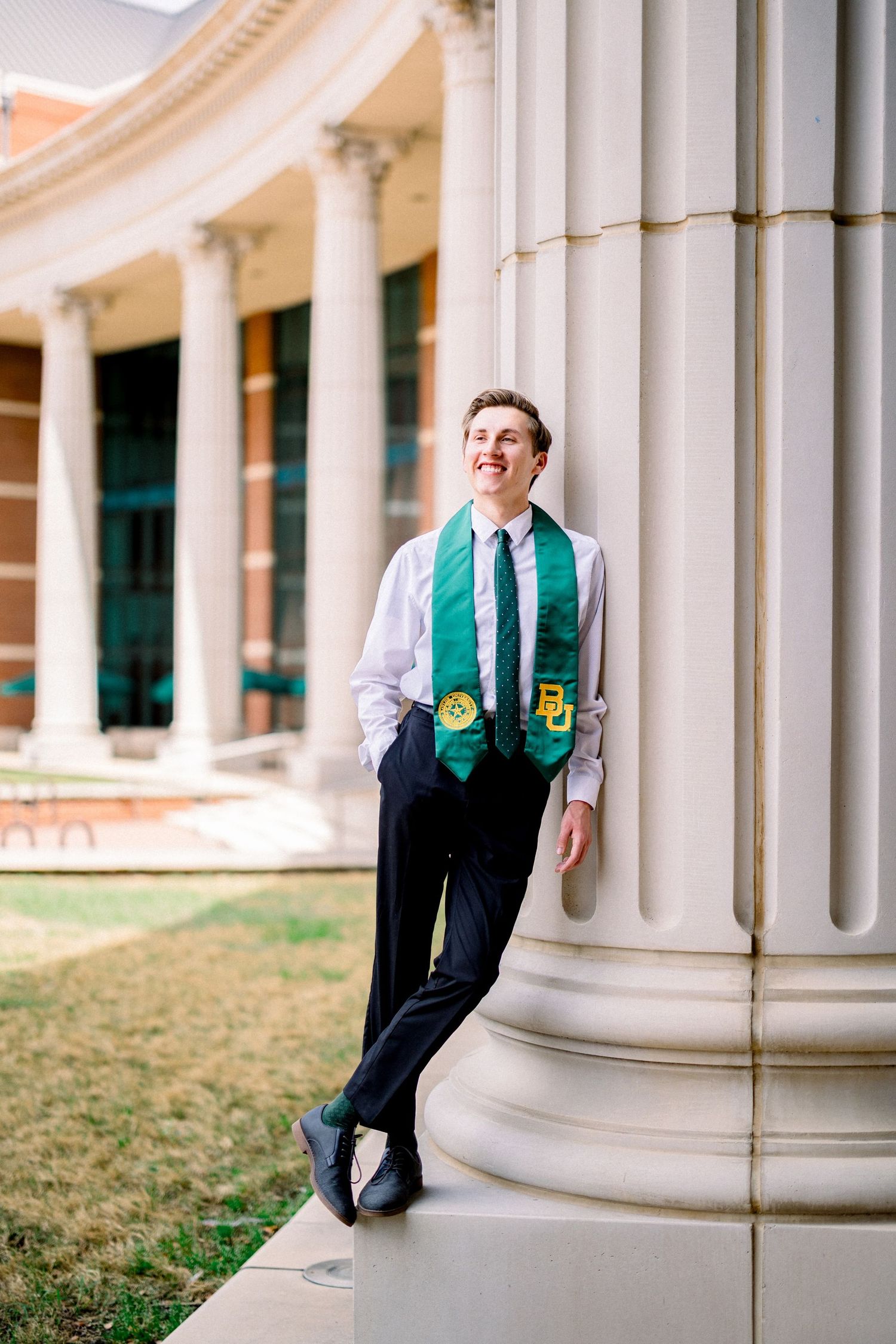 Young man in dress shirt with Baylor graduation stole leaning against white campus columns.