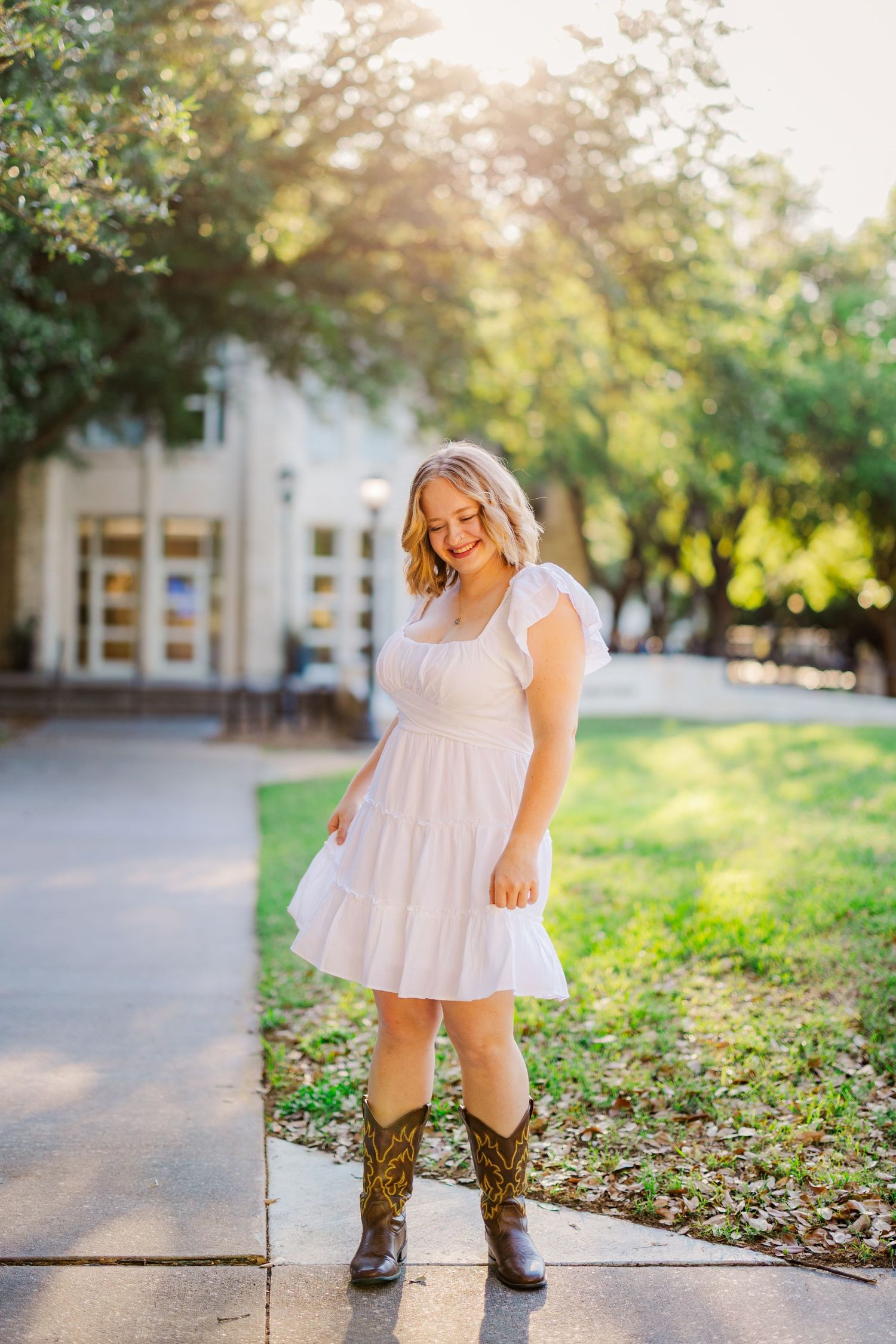 Smiling blonde teen in white dress and cowboy boots poses on a sunny campus sidewalk.