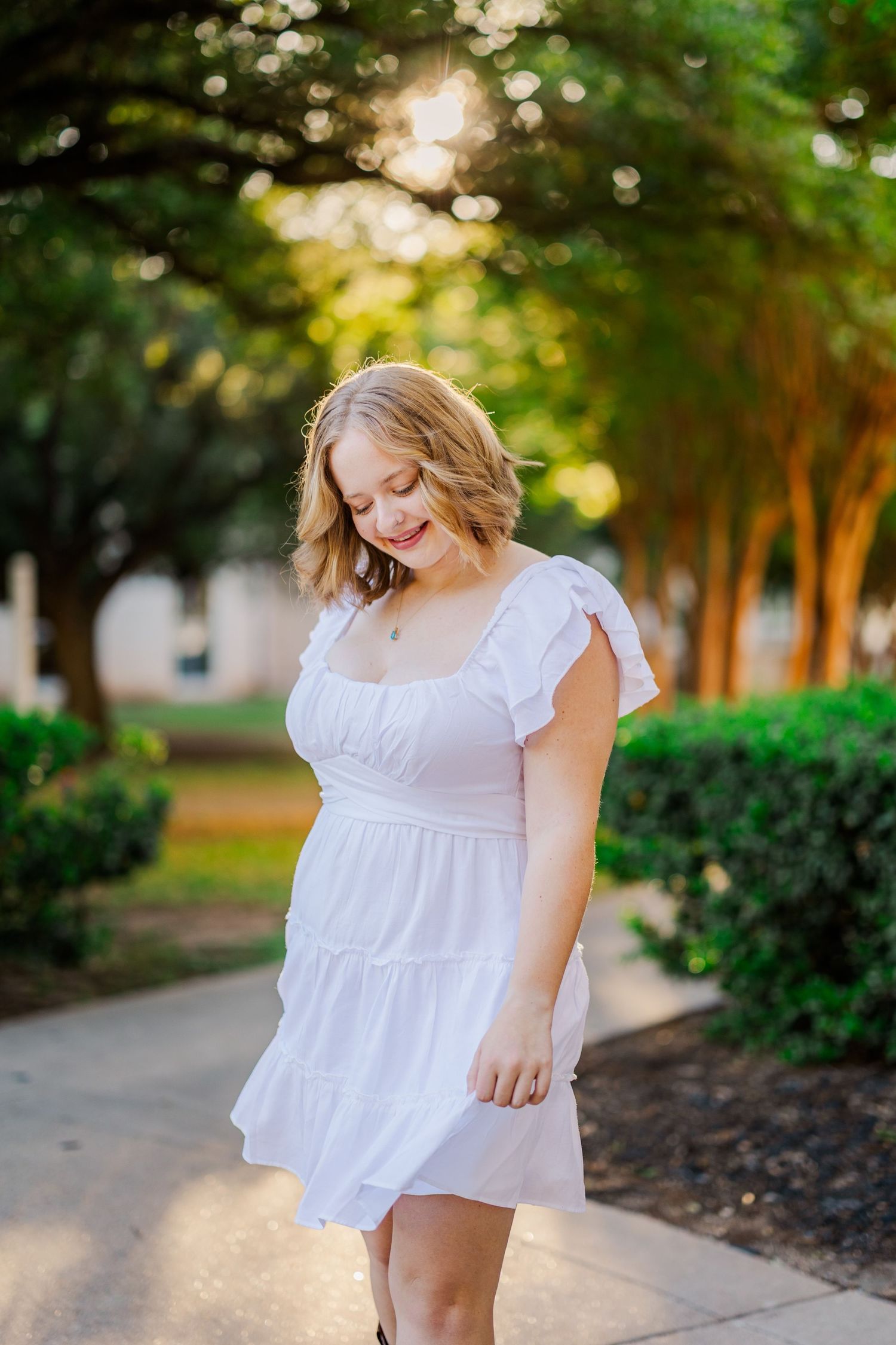 Young blonde woman in white sundress smiling in tree-lined pathway during golden hour senior photo.
