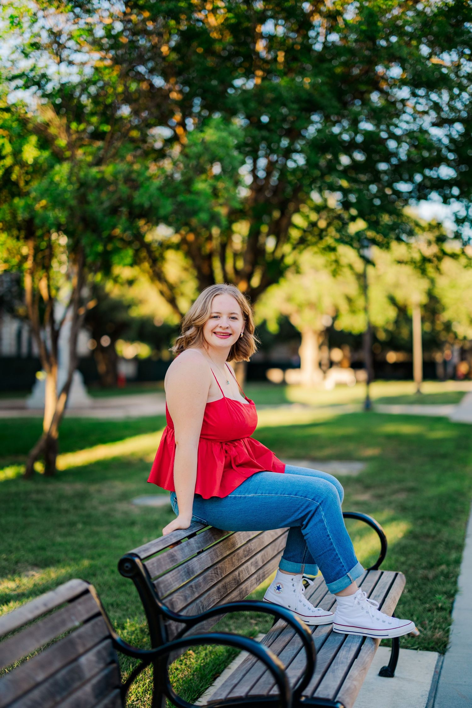 Smiling young woman in red top and jeans sits on a park bench surrounded by lush green trees.