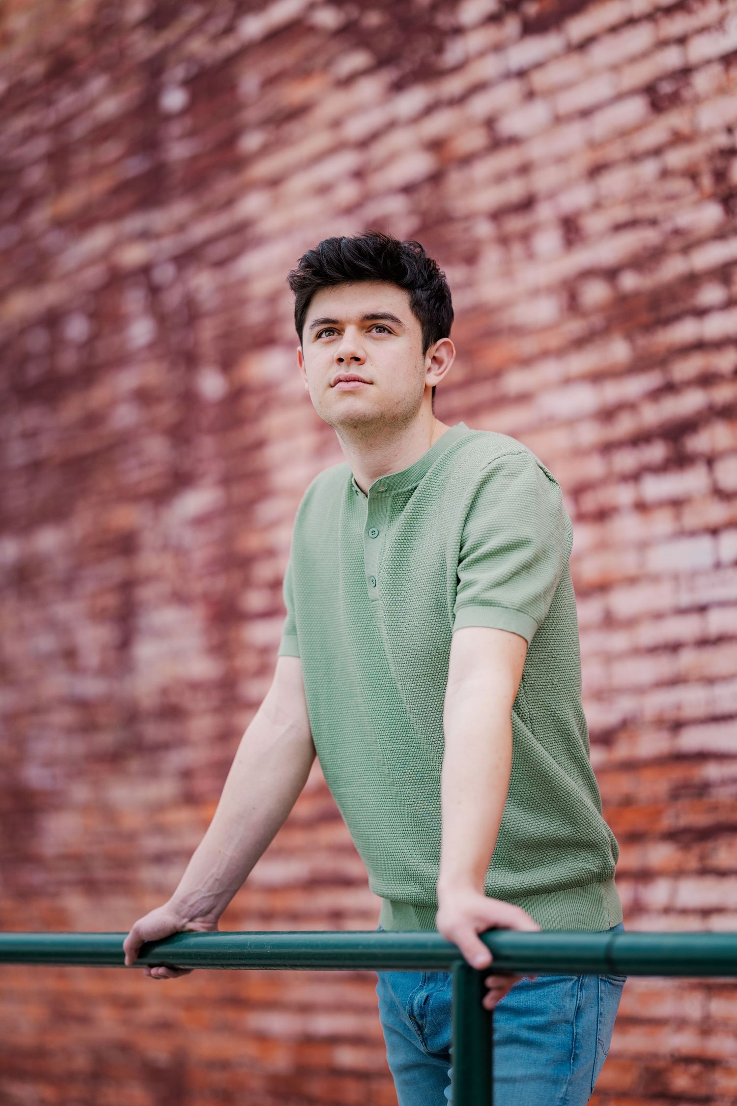 Handsome young man in green henley leans on metal rail against rustic red brick wall outdoors.