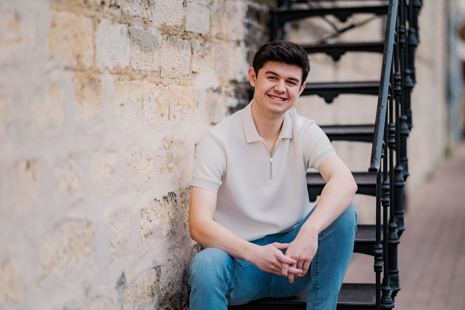 Smiling young man in beige polo sitting on stone steps beside a brick wall.