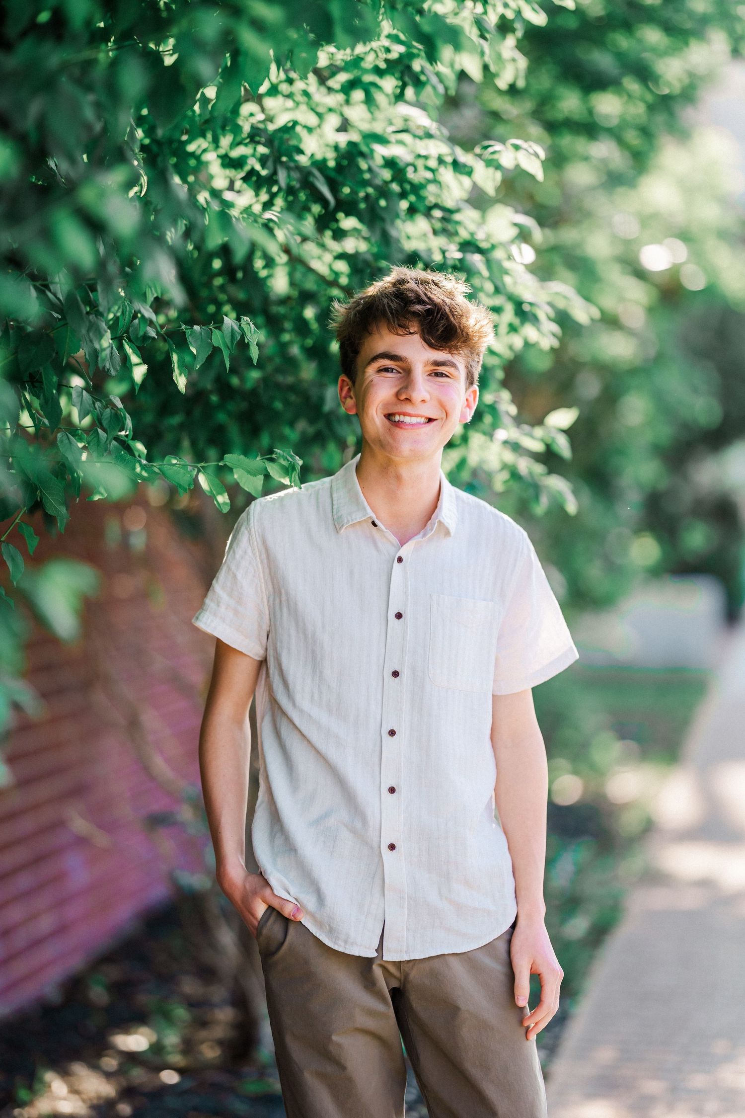 Smiling teenage boy in white button-up shirt standing outdoors surrounded by vibrant green foliage.
