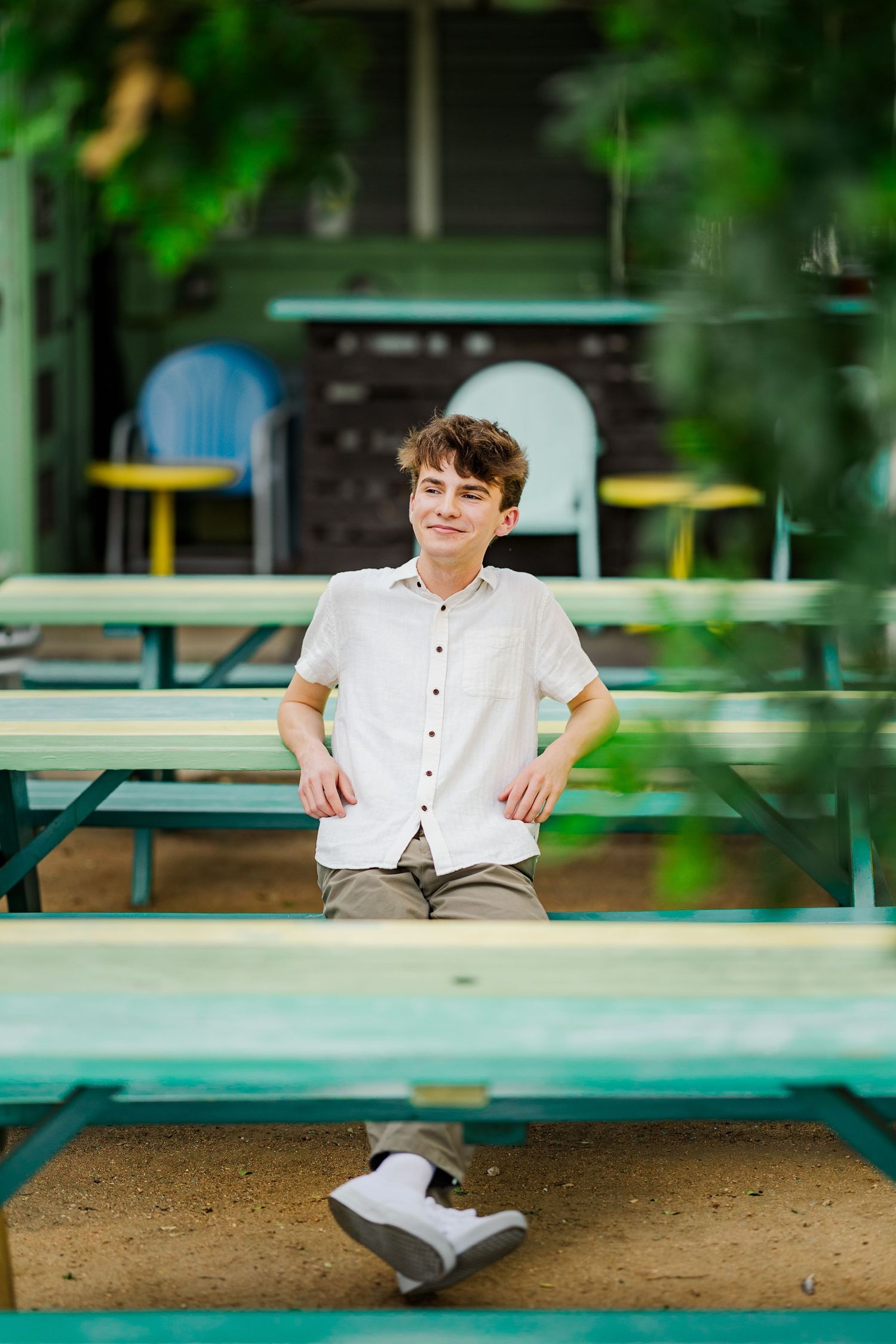 Teen boy in white shirt sits relaxed at teal picnic table in outdoor patio area with green foliage backdrop.