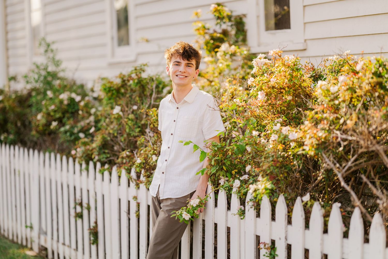 Smiling teenage boy in white shirt stands by white picket fence with blooming bushes outdoors.