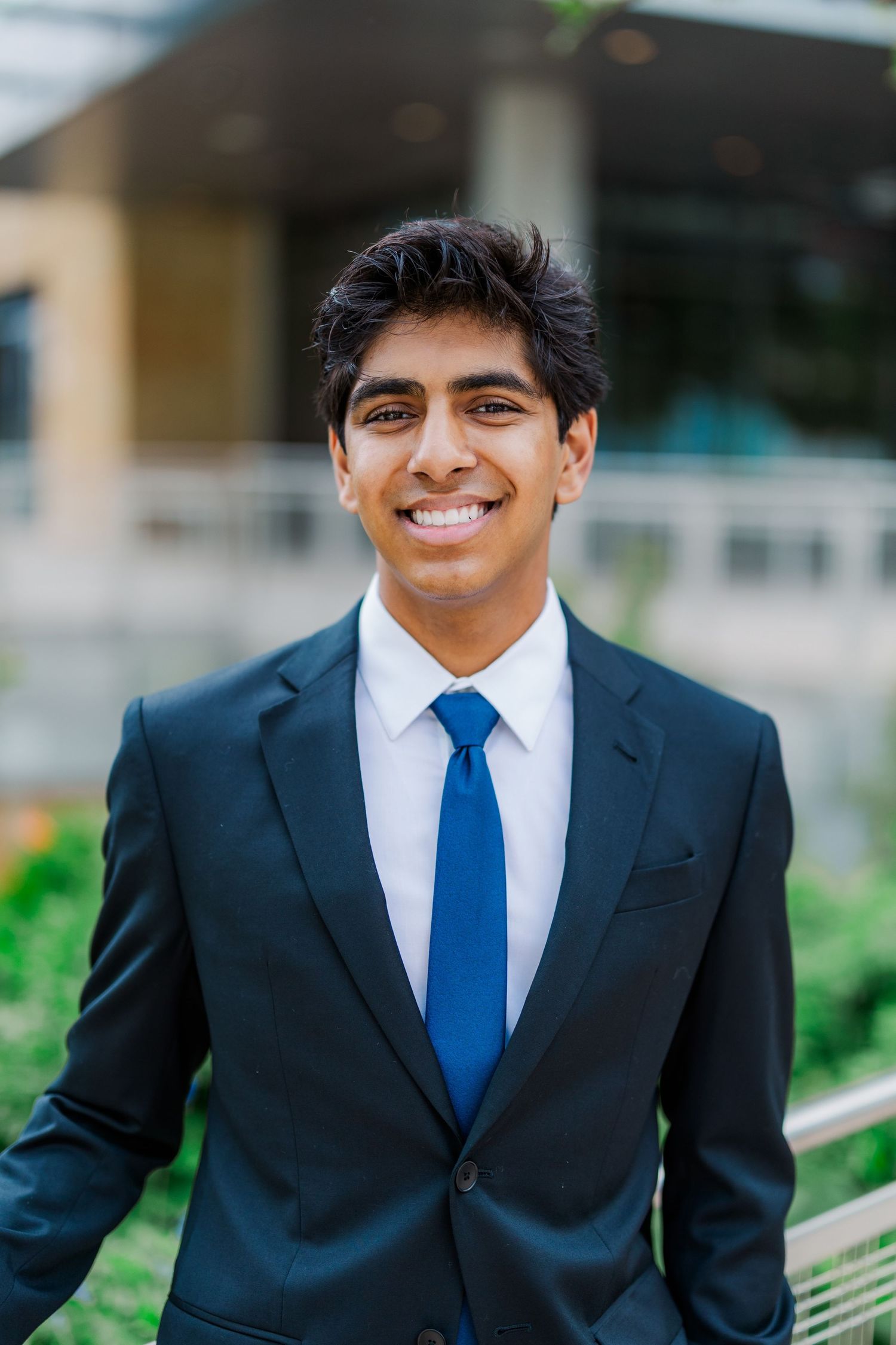 Young man in black suit and blue tie smiling confidently in modern building exterior.