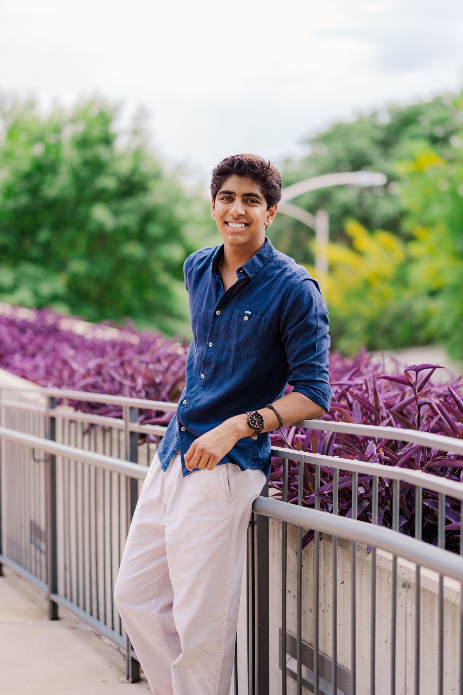 Smiling teen boy in blue linen shirt leans on metal railing with purple flower garden background.