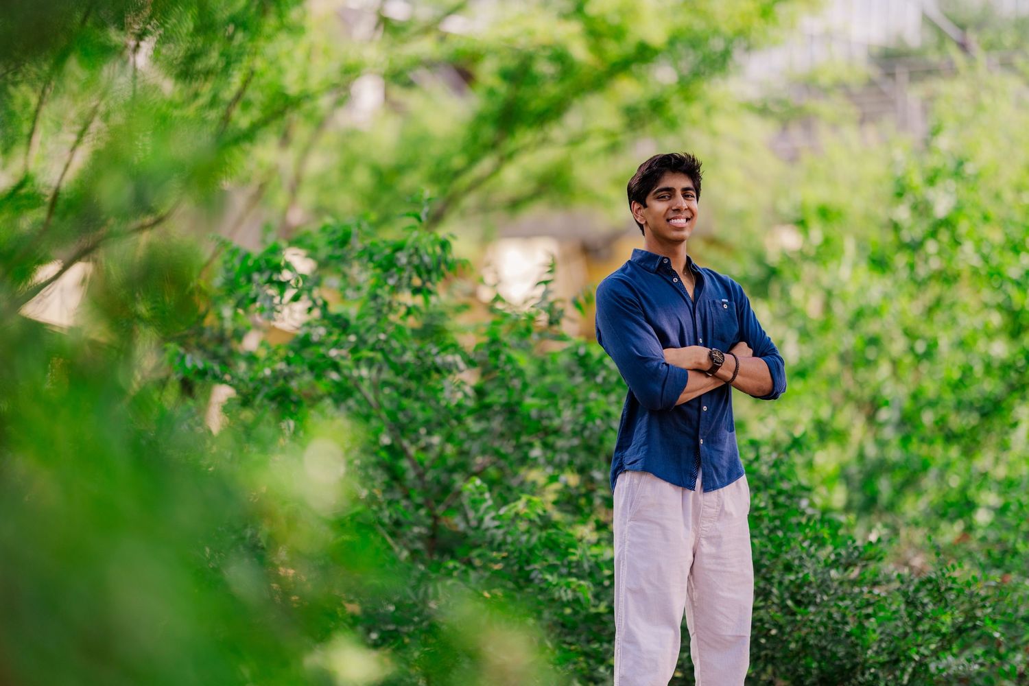 Young man in navy shirt and white pants smiling with arms crossed in lush green garden.